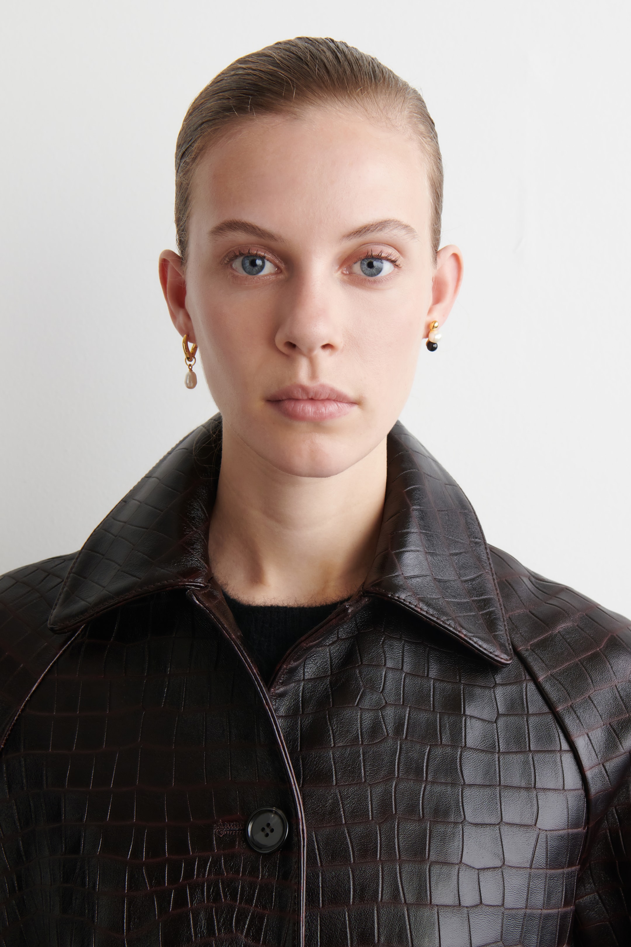 Grösseres Bild ansehen: Eine dunkelbraune Jacke mit einer Krokodilprägung wird getragen, die einen spitzen Kragen und einen einzelnen sichtbaren Knopfverschluss an der Vorderseite aufweist. Zwei unterschiedliche Ohrringe werden ebenfalls getragen: ein goldfarbener Reifen mit einer leichten tropfenförmigen Perle am linken Ohr und ein goldfarbener Ohrring mit gestapelten weißen und schwarzen Perlen am rechten Ohr.