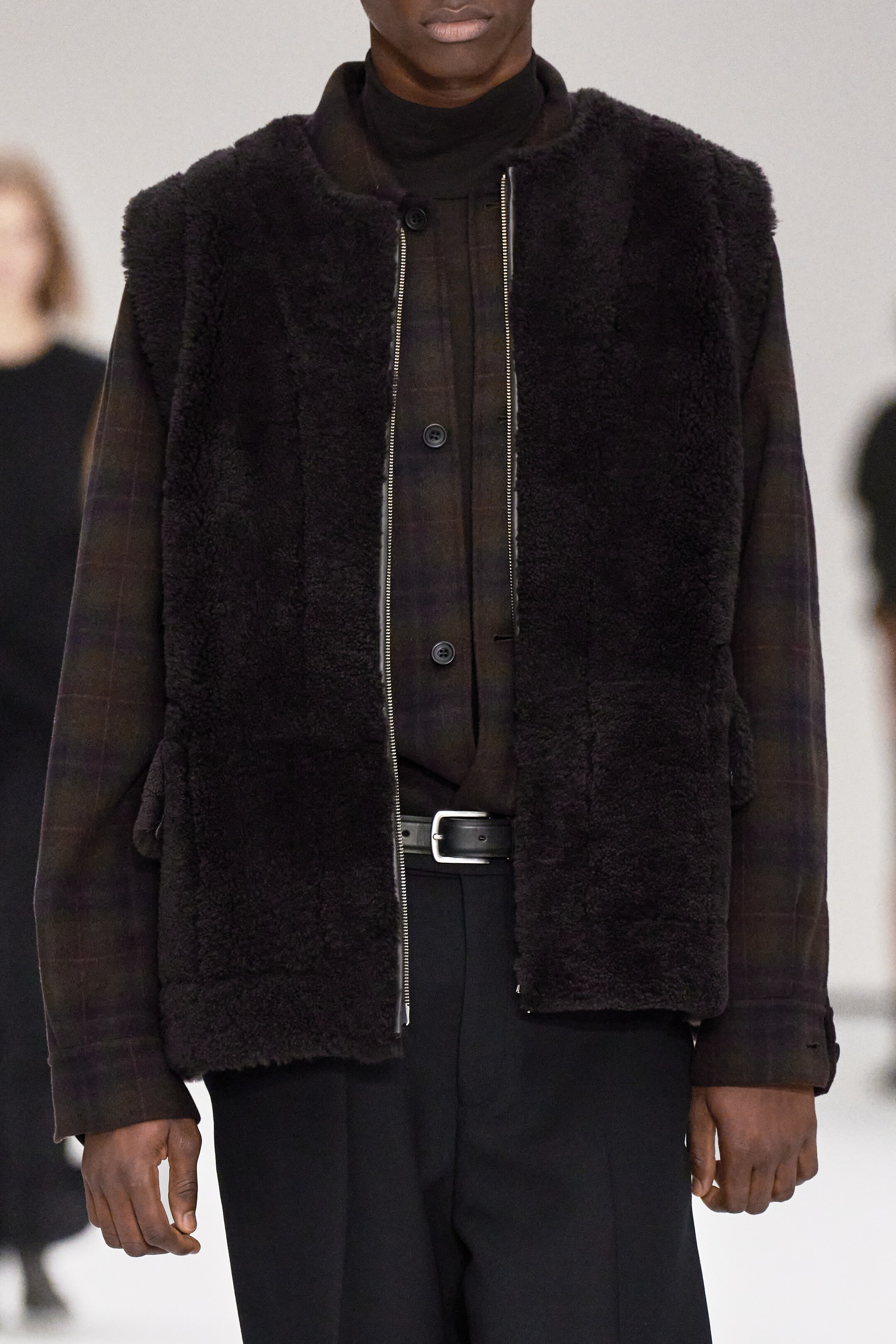 Größeres Bild anzeigen: Eine dunkelbraune, strukturierte Lammfellweste wird frontseitig getragen und zeichnet sich durch einen markanten silberfarbenen, doppelendigen Reißverschluss und sichtbare seitliche Aufgesetzte Taschen aus. Sie ist über einem dunkel karierten Overshirt mit geknöpften Manschetten und einem schwarzen Rollkragenpullover geschichtet.