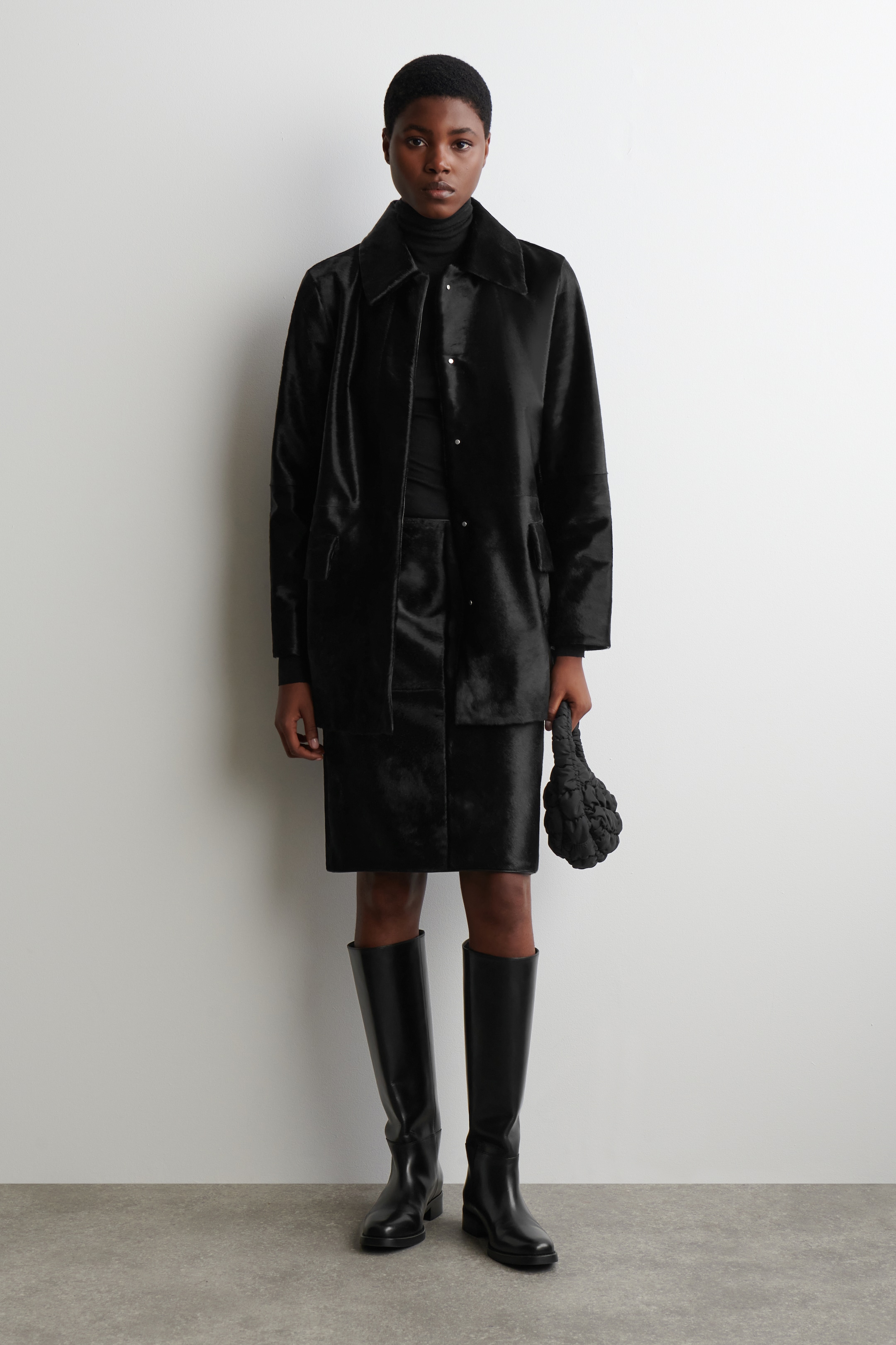 Ingrandisci l'immagine: Un ensemble nero viene presentato in una vista frontale, comprendente un cappotto e una gonna coordinata al ginocchio realizzati in un materiale a pelo corto. Il cappotto, con un colletto classico, chiusura frontale con bottoni e due tasche applicate anteriori, è stratificato su un top a collo alto nero. Il look è completato da stivali neri al ginocchio e una borsa nera testurizzata a mano.