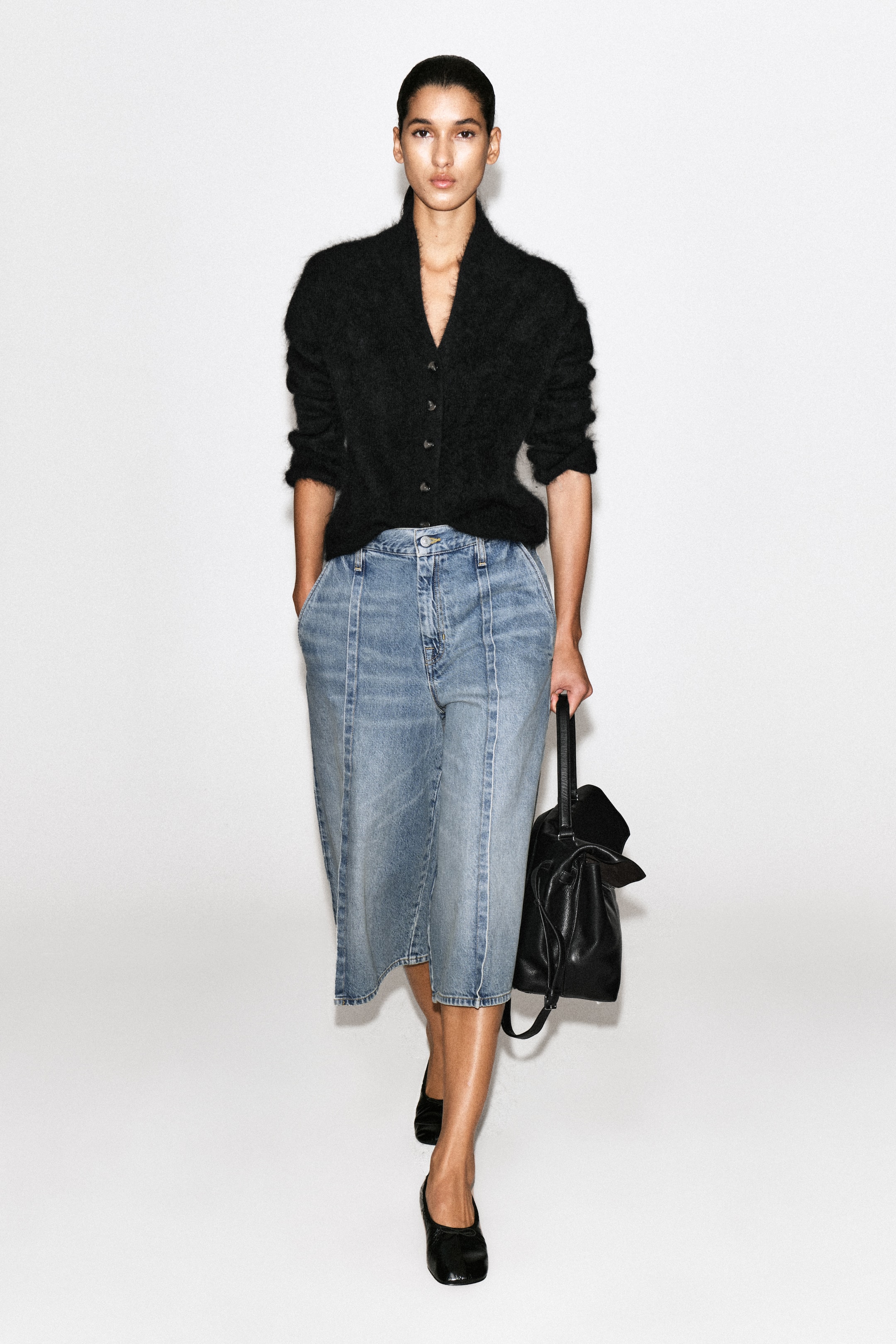 Vezi imaginea mai mare: Aceste culottes din denim albastru deschis, prezentate din față, au o croială largă, scurtată, cu cusături verticale bine definite, de tip pintuck, care coboară pe fiecare picior. Un cardigan negru, pufos, este introdus în talie, evidențiind structura cu talie înaltă și închidere cu nasturi.