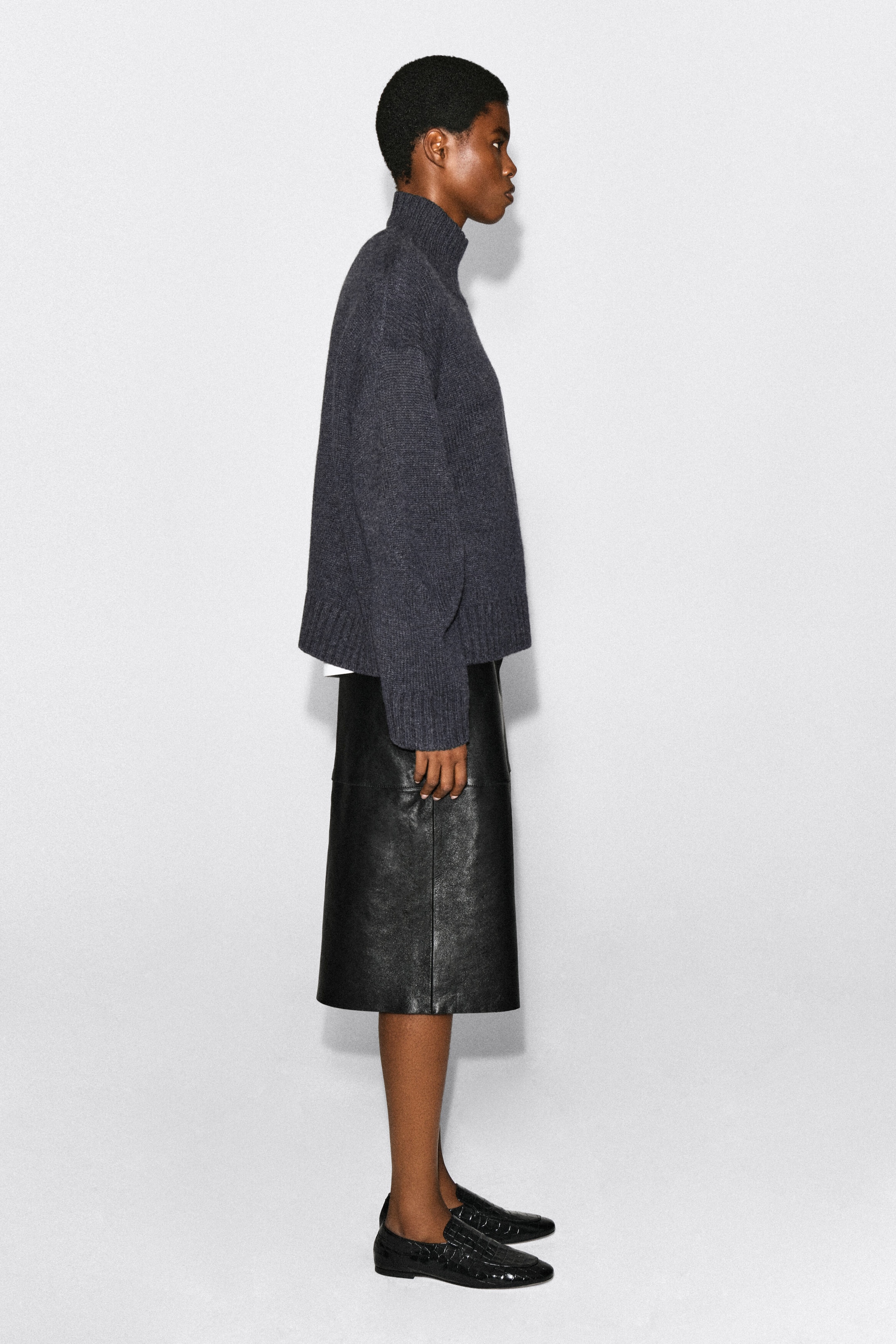 The model is wearing a chunky, dark grey cashmere funnel-neck jumper, shown in side profile. The relaxed-fit jumper features dropped shoulders and ribbed detailing on the collar, cuffs and hem, and is styled with a black leather skirt. The model is wearing a chunky, dark grey cashmere funnel-neck jumper, shown in side profile. The relaxed-fit jumper features dropped shoulders and ribbed detailing on the collar, cuffs and hem, and is styled with a black leather skirt.
