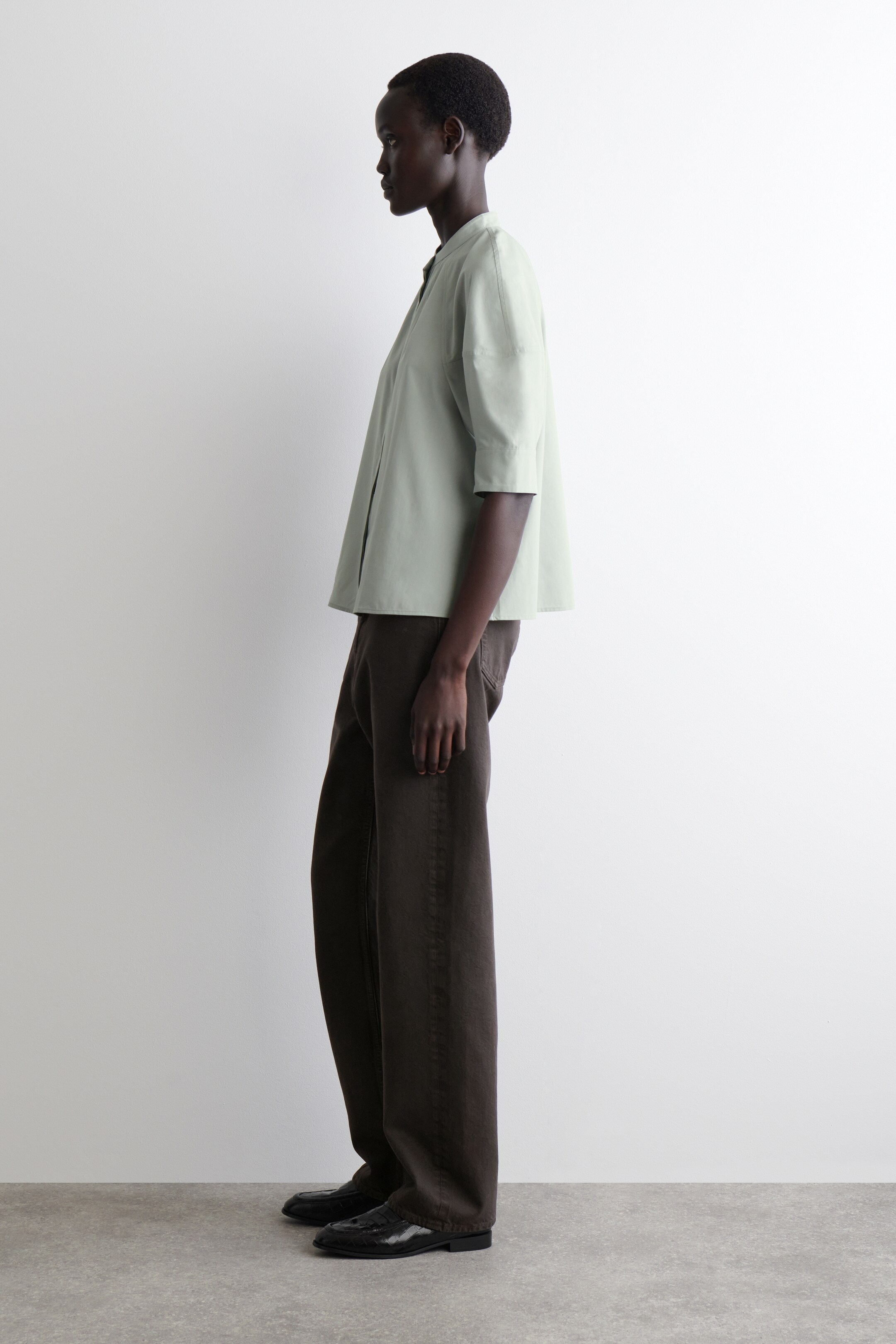 Grotere afbeelding bekijken: Een lichtgroene blouse wordt gemodelleerd met de voorkant naar links. De blouse heeft een afgeronde grandad-kraag, volumineuze raglanmouwen tot aan de elleboog en een subtiel uitlopende, normale pasvorm.
