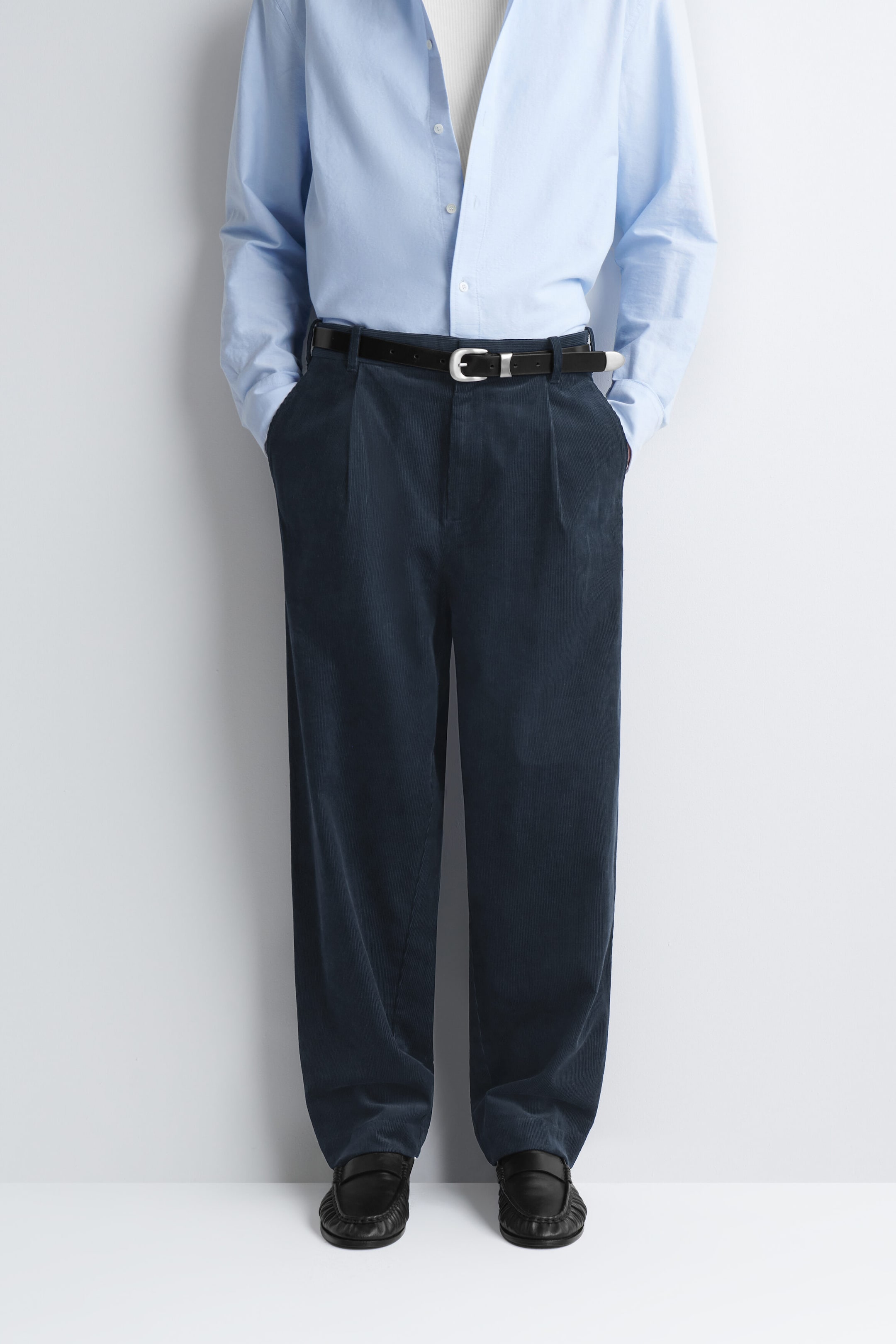 Dark navy corduroy relaxed-fit tapered pants with front pleats are worn by the model, with hands resting in the side pockets. The pants are styled with a black leather belt, a light blue long-sleeved shirt, and black leather loafers. Dark navy corduroy relaxed-fit tapered pants with front pleats are worn by the model, with hands resting in the side pockets. The pants are styled with a black leather belt, a light blue long-sleeved shirt, and black leather loafers.