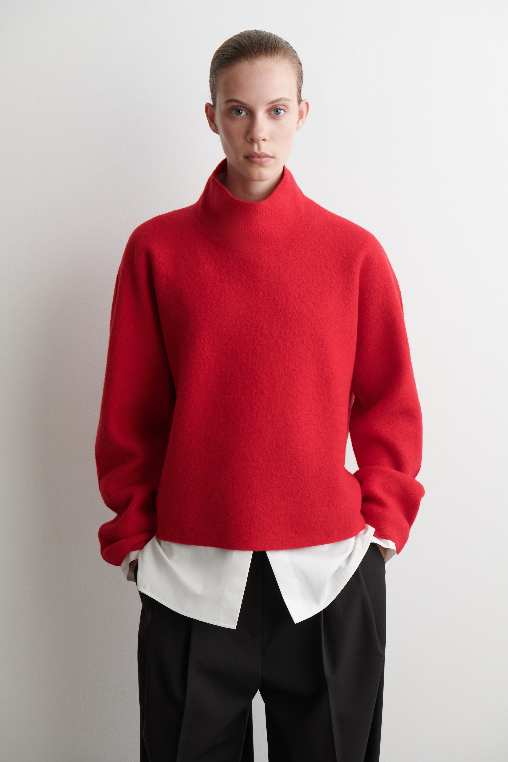 Agrandir l'image: Un pull à col montant rouge est présenté de face, avec une coupe ample et carrée et un tissu en tricot brossé texturé. Une chemise blanche à col est visiblement superposée en dessous, s'étendant sous l'ourlet droit du pull et émergeant de ses larges ouvertures de manches.