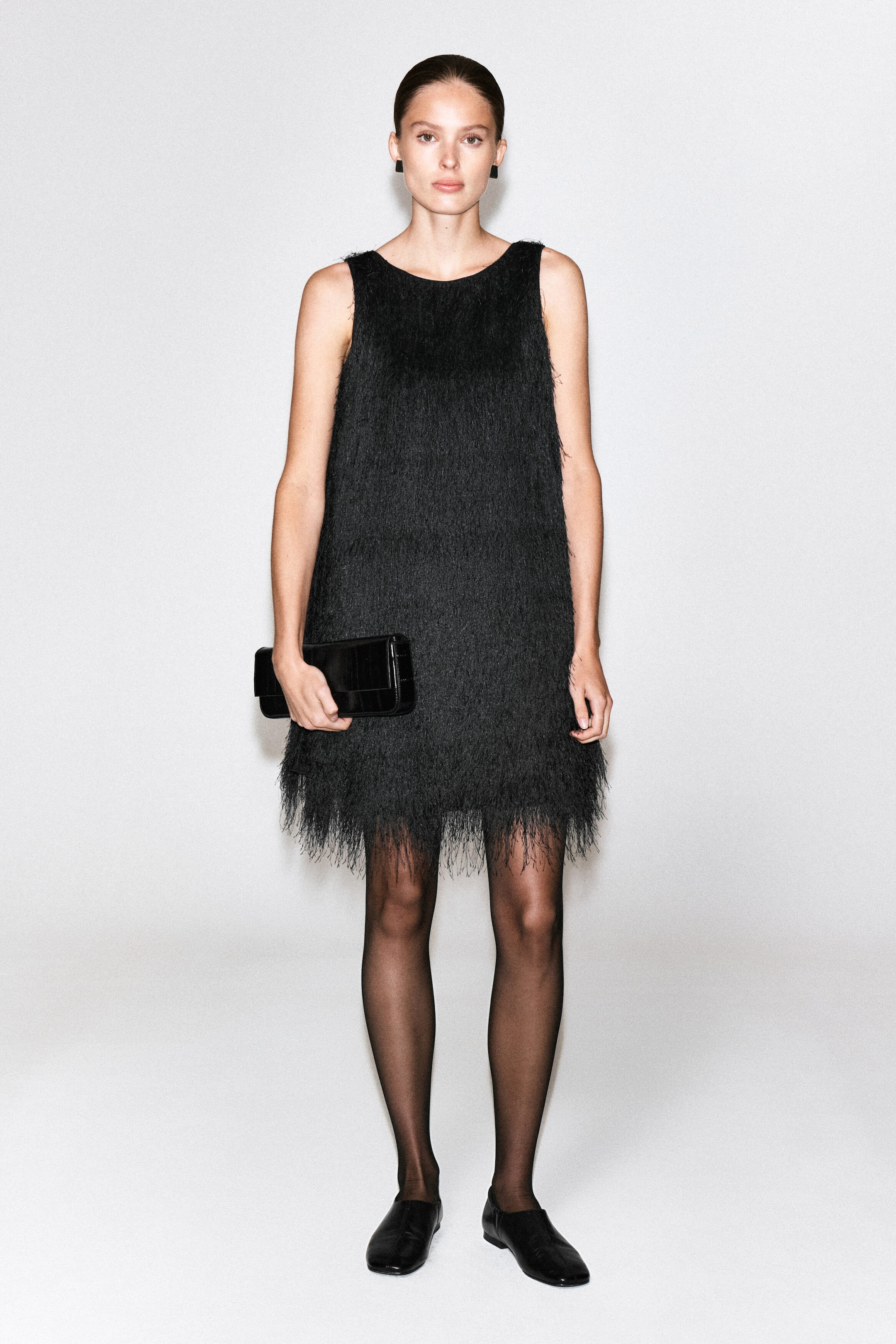 Ein schwarzes Mini-Partykleid mit durchgehender feiner Fransenstruktur, ärmellosem Schnitt und rundem Ausschnitt wird von dem Model getragen. Das Kleid ist mit transparenten schwarzen Strumpfhosen und flachen Schuhen kombiniert, während das Model eine schwarze Clutch in der Hand hält. Ein schwarzes Mini-Partykleid mit durchgehender feiner Fransenstruktur, ärmellosem Schnitt und rundem Ausschnitt wird von dem Model getragen. Das Kleid ist mit transparenten schwarzen Strumpfhosen und flachen Schuhen kombiniert, während das Model eine schwarze Clutch in der Hand hält.