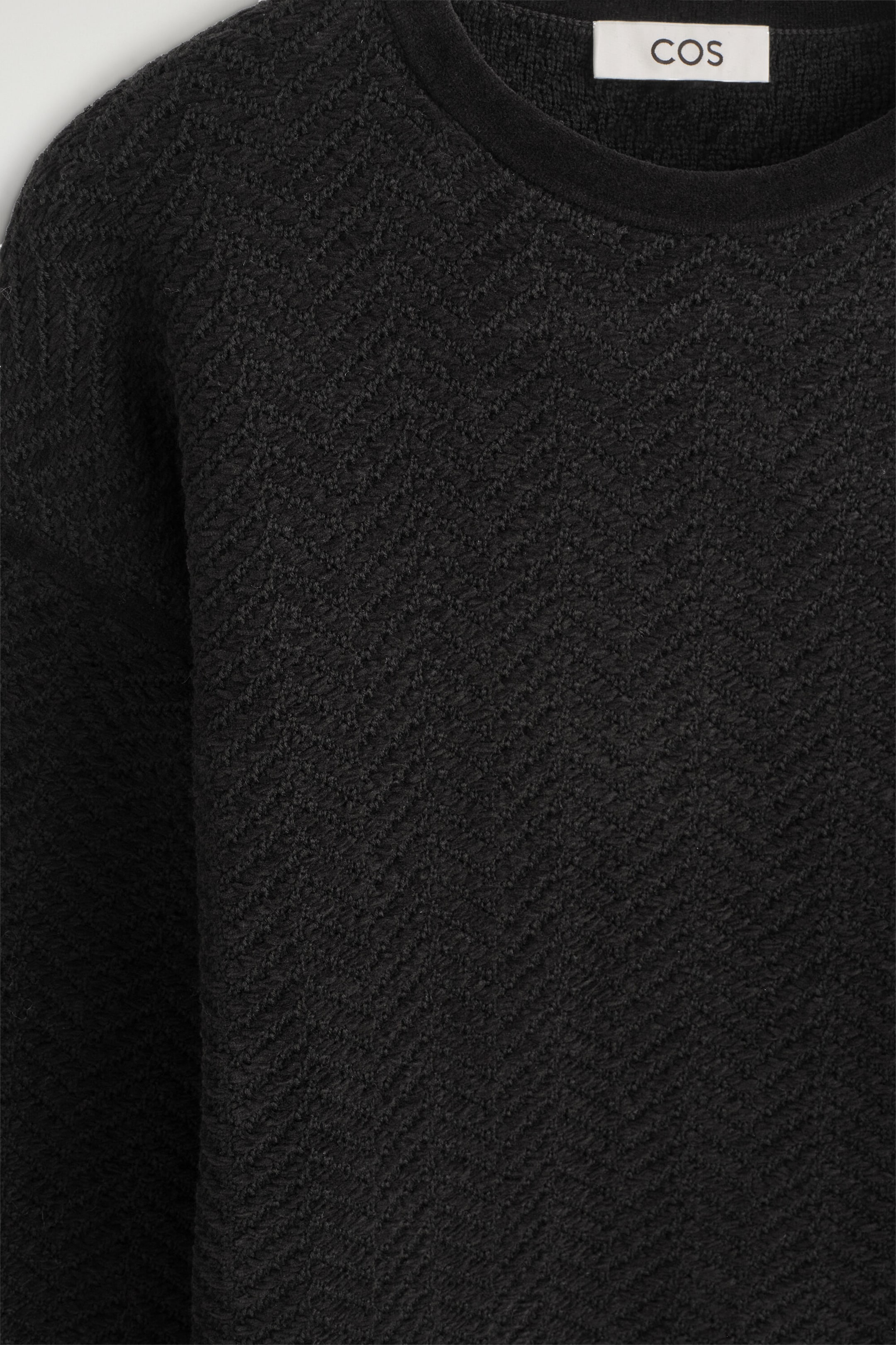 This close-up front view shows the black merino wool chenille crew-neck sweater, highlighting its textured, chevron-patterned knit. The sweater features a smooth, finely ribbed crew neckline and relaxed dropped shoulders. This close-up front view shows the black merino wool chenille crew-neck sweater, highlighting its textured, chevron-patterned knit. The sweater features a smooth, finely ribbed crew neckline and relaxed dropped shoulders.
