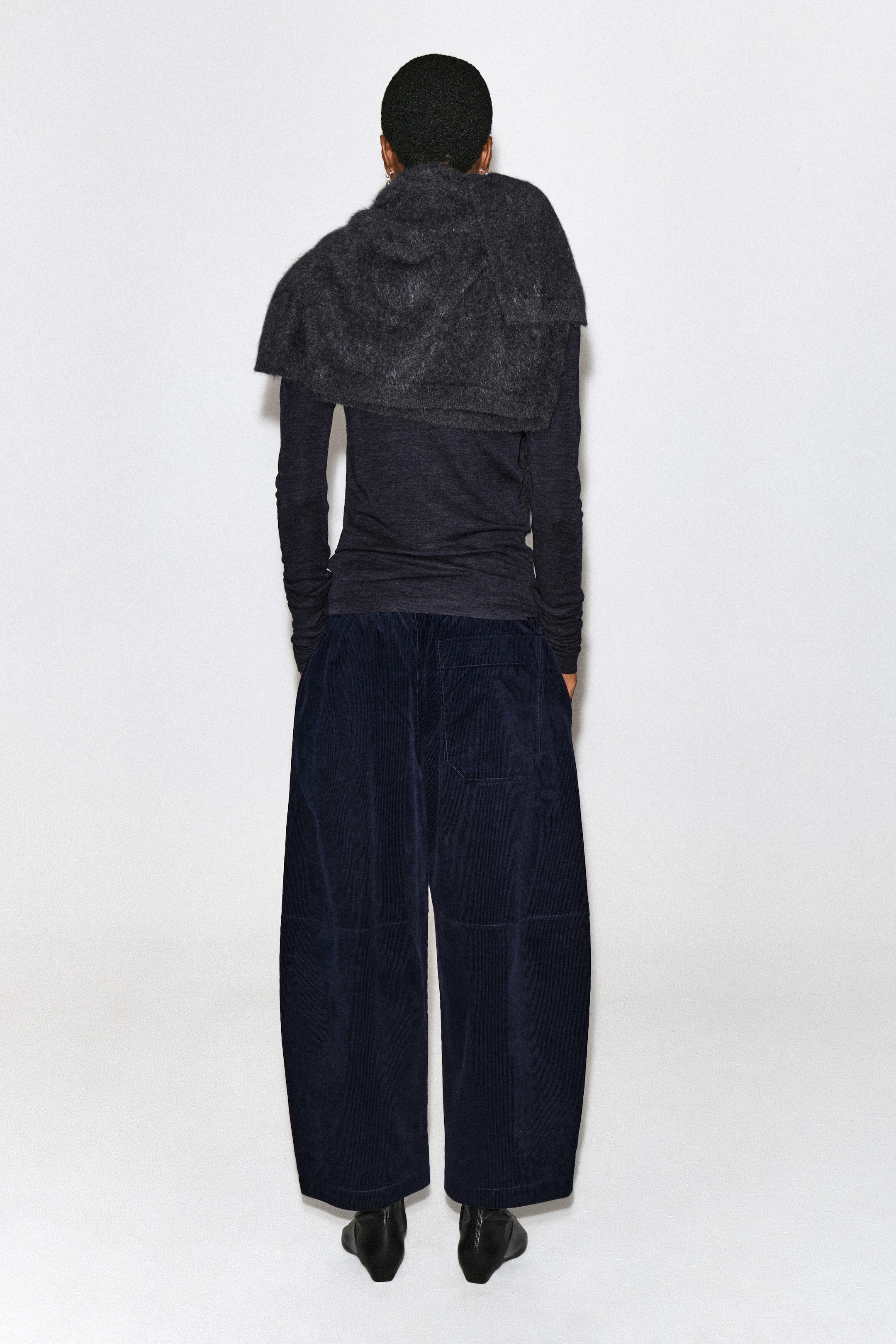 Corduroy barrel-leg pants in dark navy blue are worn by the model, who is standing with their back to the camera. The pants feature a wide, curved silhouette, horizontal knee seams, and a single back patch pocket, styled with a dark long-sleeve top, a textured gray knit scarf, and black boots. Corduroy barrel-leg pants in dark navy blue are worn by the model, who is standing with their back to the camera. The pants feature a wide, curved silhouette, horizontal knee seams, and a single back patch pocket, styled with a dark long-sleeve top, a textured gray knit scarf, and black boots.