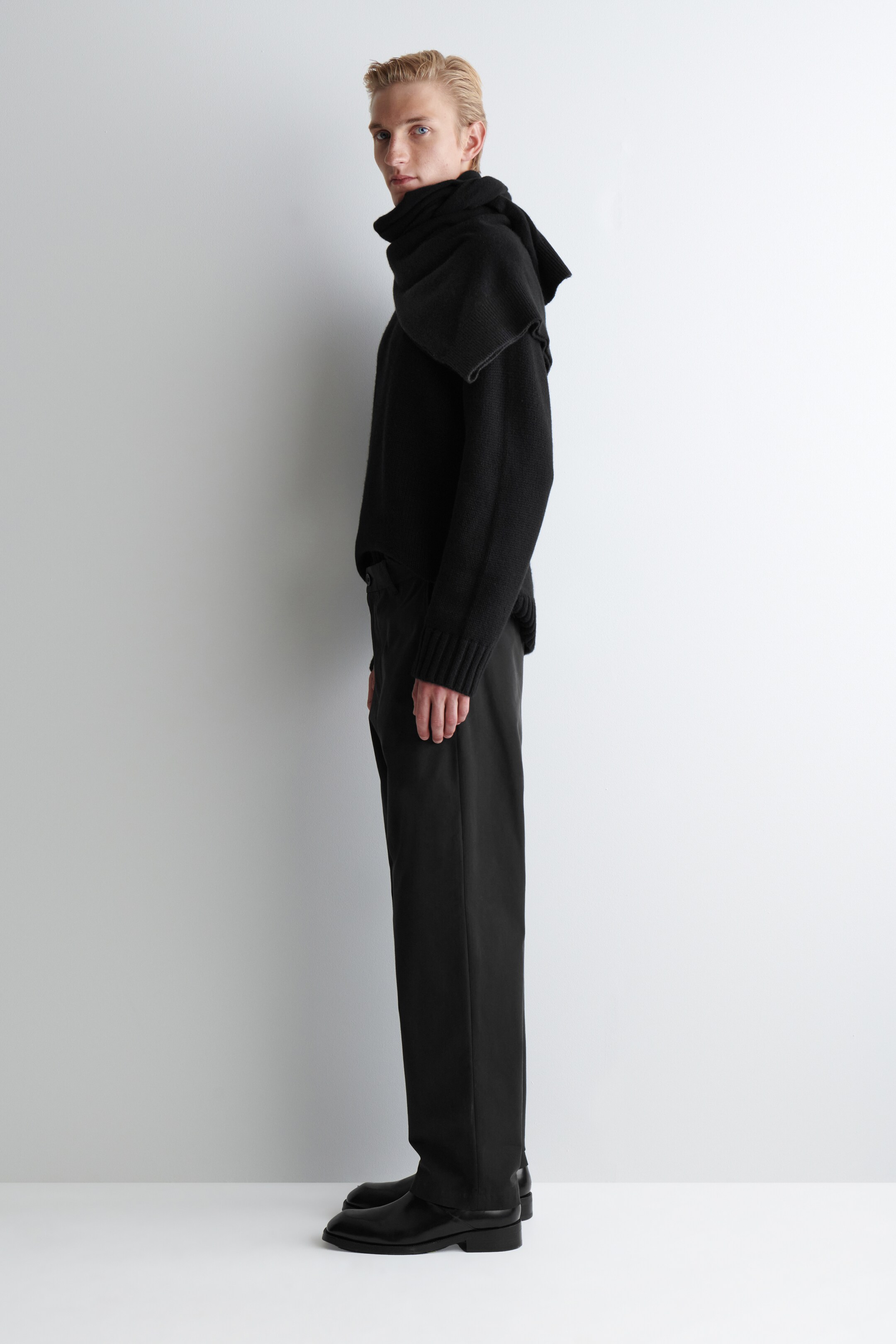 Visualizza immagine più grande: Un top a maglia nero a maniche lunghe, caratterizzato da un collo alto e voluminoso che si drappeggia sulle spalle e polsini a coste, è mostrato in un profilo laterale. È abbinato a pantaloni neri a vestibilità rilassata e scarpe eleganti in pelle nera.