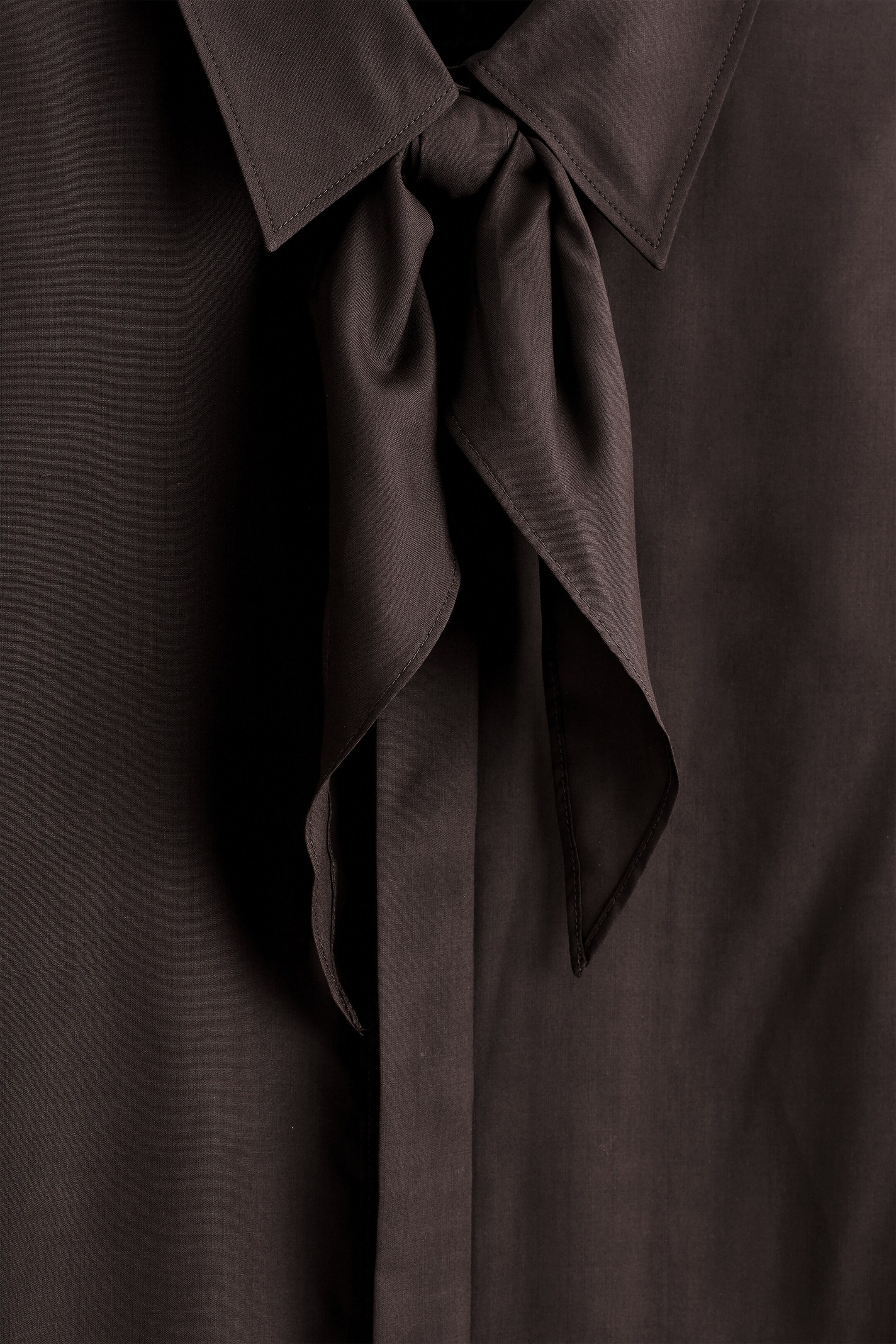 Agrandir l'image: Une chemise en soie de ton marron est présentée de face, avec un col pointu et une écharpe ton sur ton nouée au niveau de l'encolure. Le tissu lustré présente des détails de couture fins sur le col et des plis subtils sur le corps de la chemise.