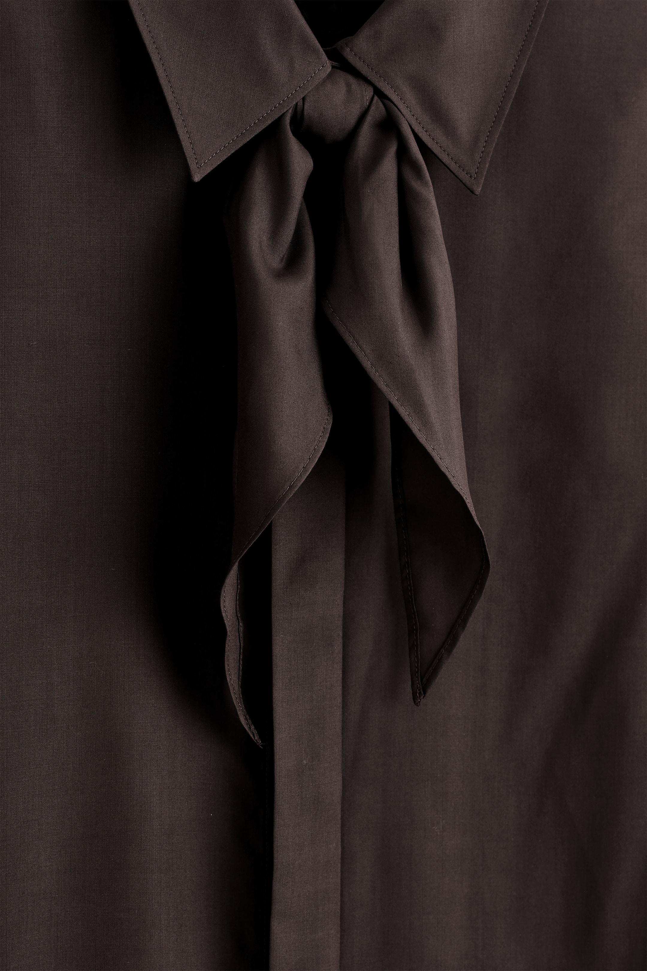 Ver imagen más grande: Una camisa de seda de tono castaño se muestra desde el frente, con un cuello puntiagudo y una bufanda tonal atada en un nudo en el escote. La tela lustrosa muestra detalles de costura fina en el cuello y pliegues sutiles en el cuerpo de la camisa.