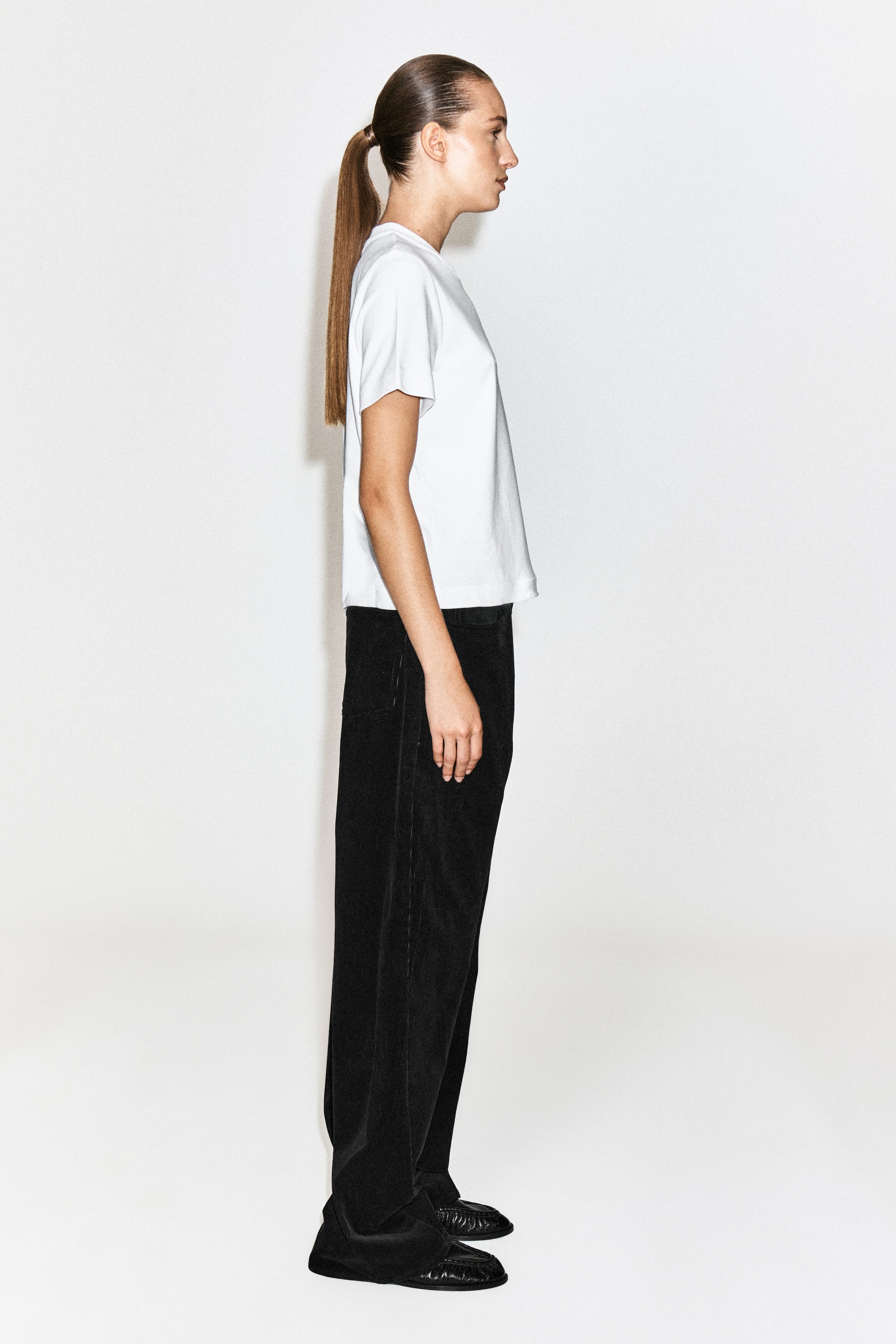 The model is wearing a white short-sleeved crew-neck T-shirt and black wide-leg trousers, shown in side profile. The T-shirt features a straight hem and relaxed fit, while the trousers have a soft texture and a loose drape.