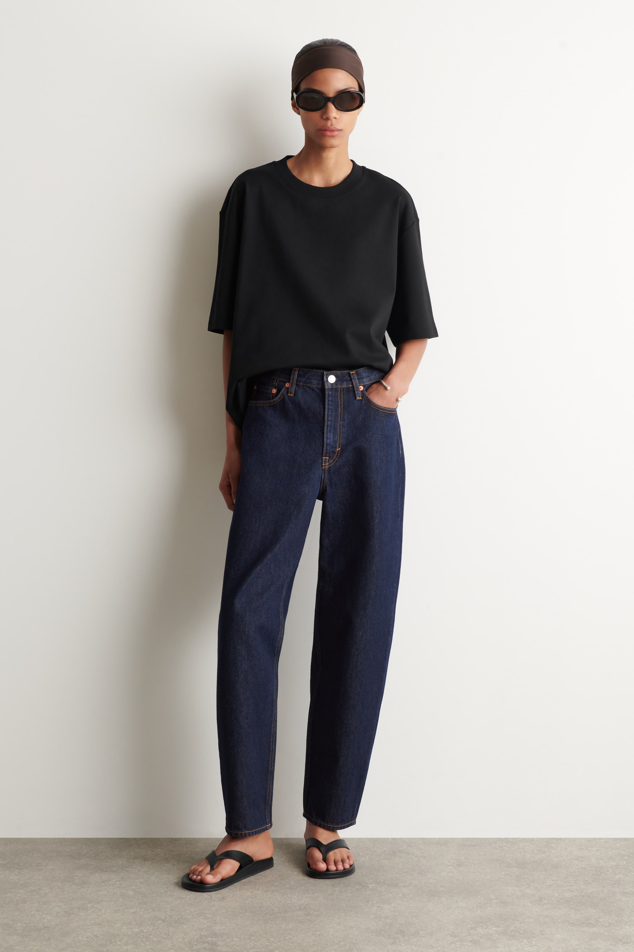 Vis større bilde: En svart t-skjorte med rund hals og korte ermer presenteres forfra, og viser et oversized draperi og er stukket inn i mørkvaskede, høytlivs jeans. Jeansene har synlige oransje sømmer og en avslappet, rett passform.