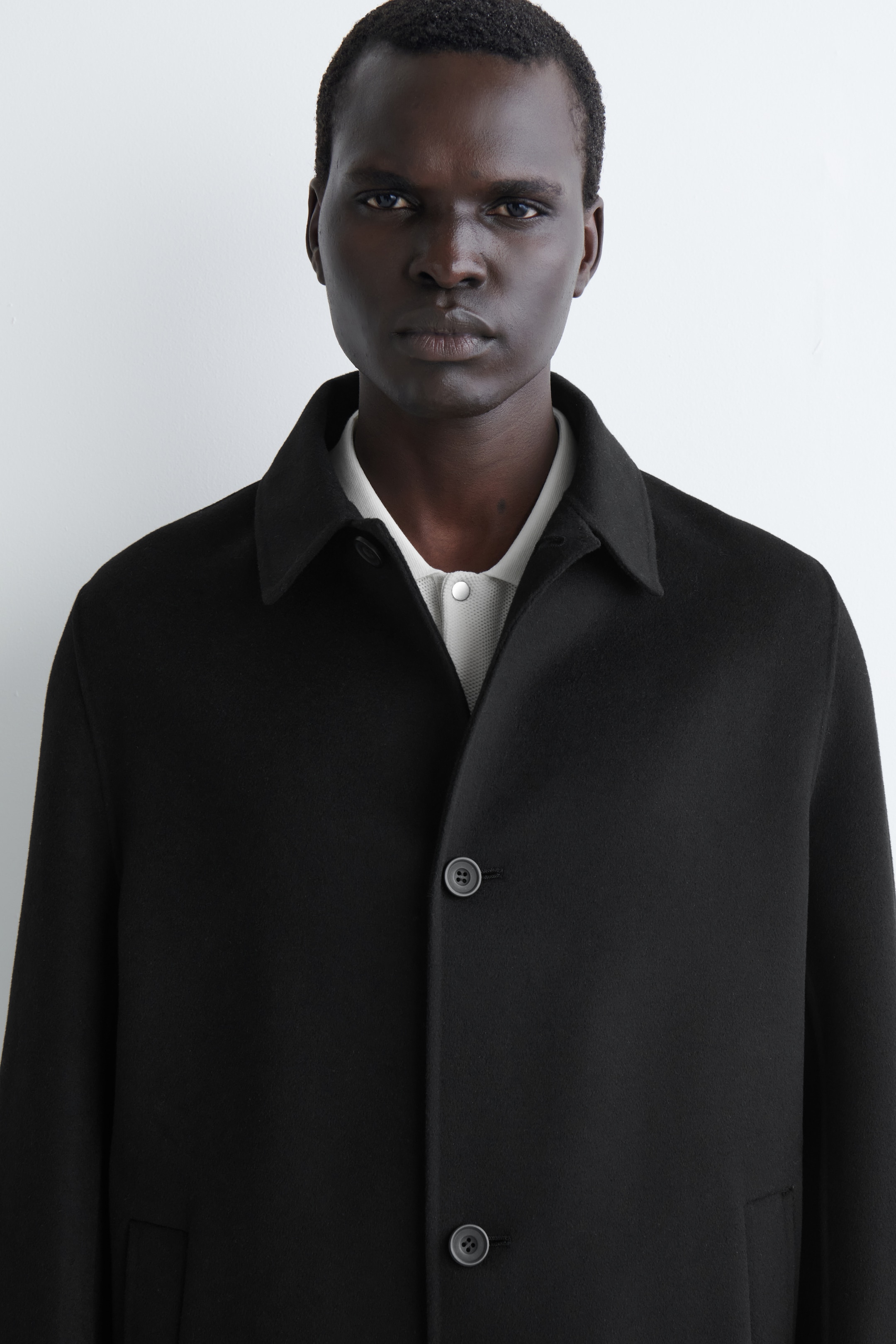 Visualizza immagine più grande: Un cappotto nero è presentato frontalmente, caratterizzato da un classico colletto a punta e una chiusura frontale con due bottoni grigio scuro visibili. Una polo di colore chiaro e con una texture particolare è indossata sotto, e una tasca a filetto è visibilmente presente in modo discreto in basso a destra.