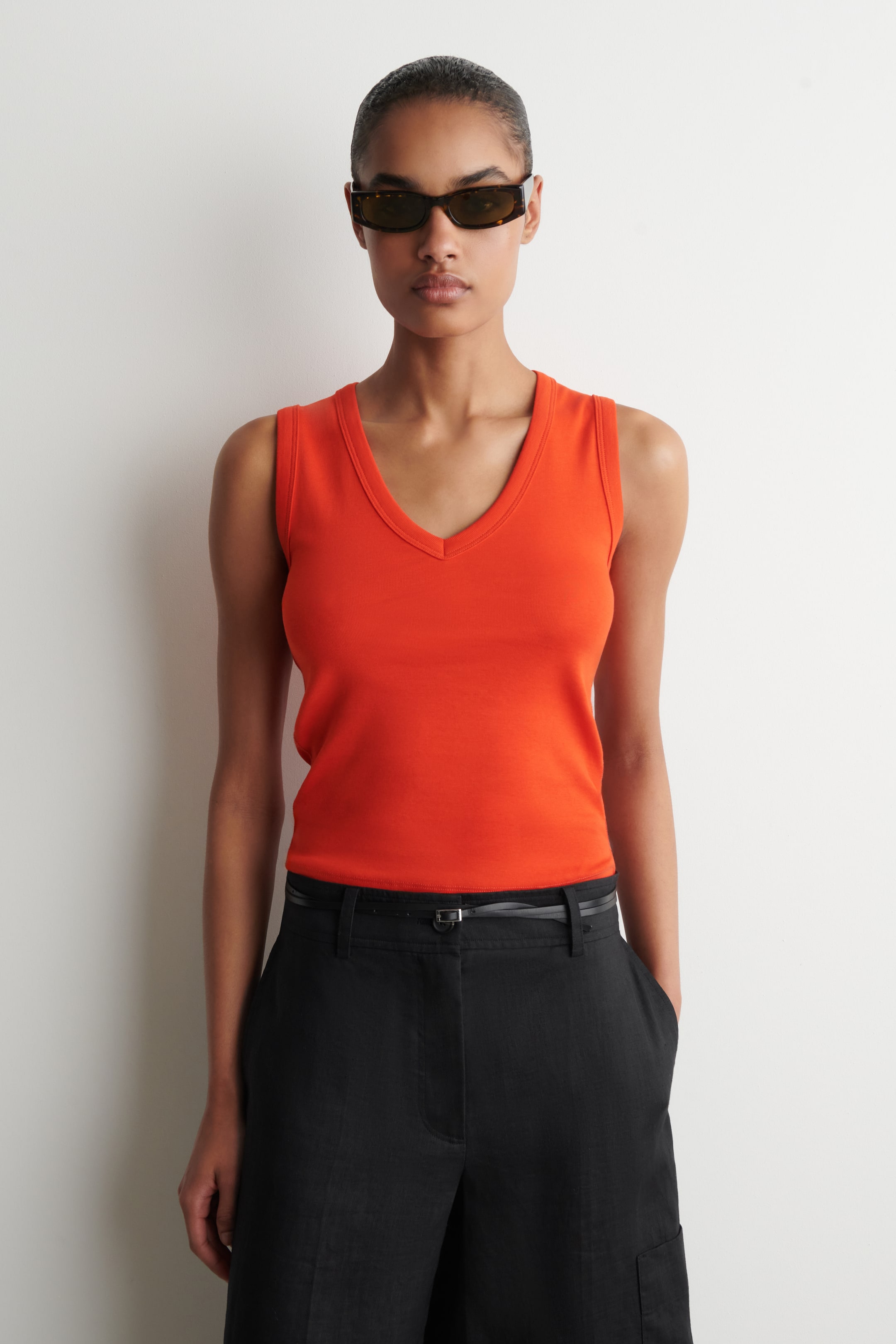 Agrandir l'image: Un haut sans manches orange avec un décolleté en V et une silhouette ajustée est porté, présenté rentré dans un pantalon noir.