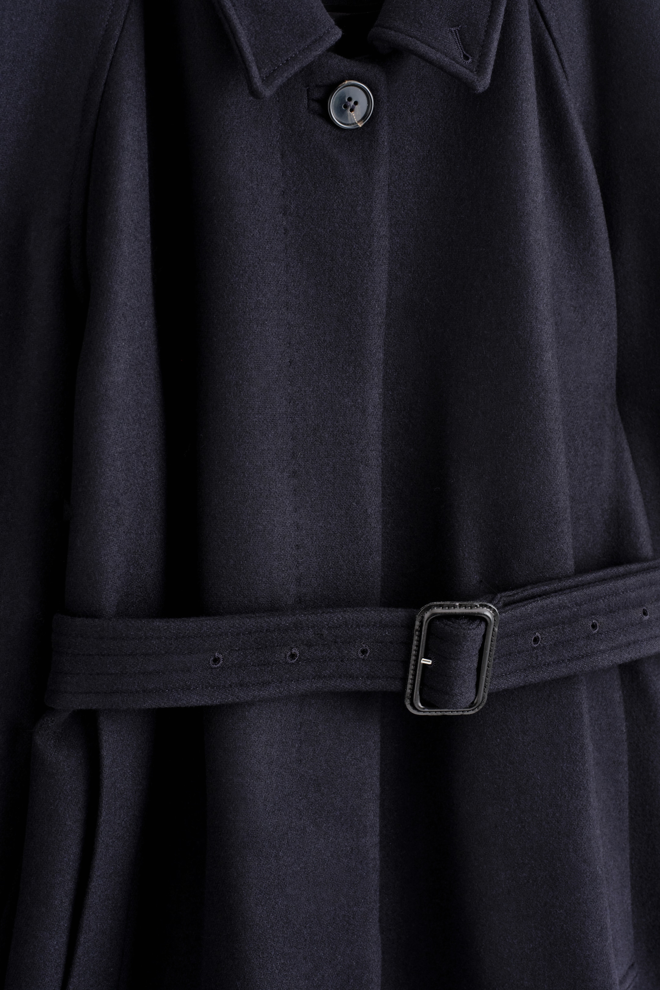 Agrandir l'image: Un manteau en laine bleu marine foncé est présenté de face, avec un col pointu et une fermeture à bouton unique visible. Une ceinture assortie avec une boucle rectangulaire foncée resserre la silhouette décontractée à la taille.
