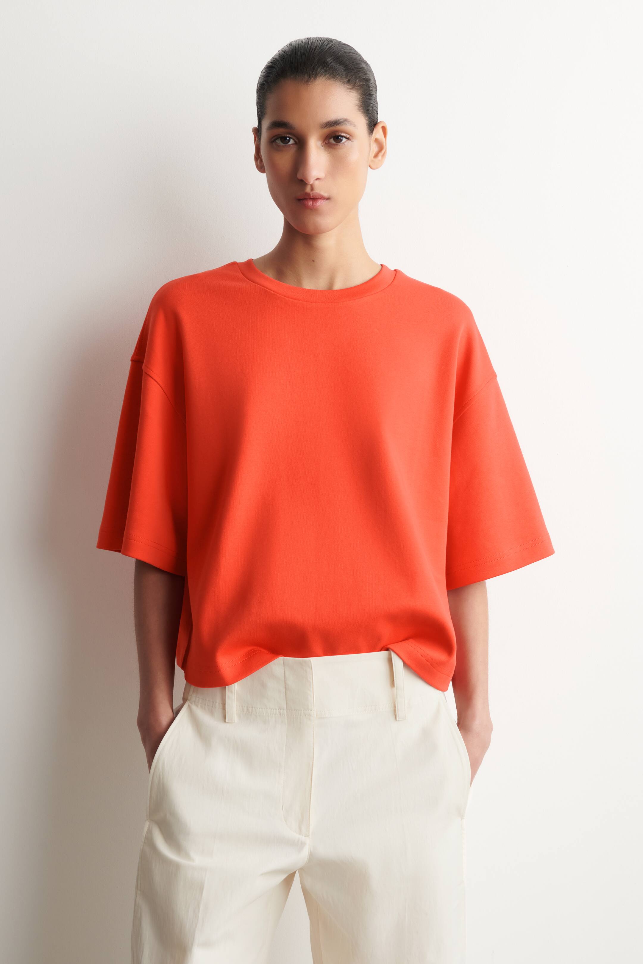 Agrandir l'image: Un t-shirt orange à col rond présente une coupe ample avec des manches courtes et larges. Il est porté avec un pantalon taille haute de couleur crème, doté de poches latérales visibles et de passants pour ceinture.