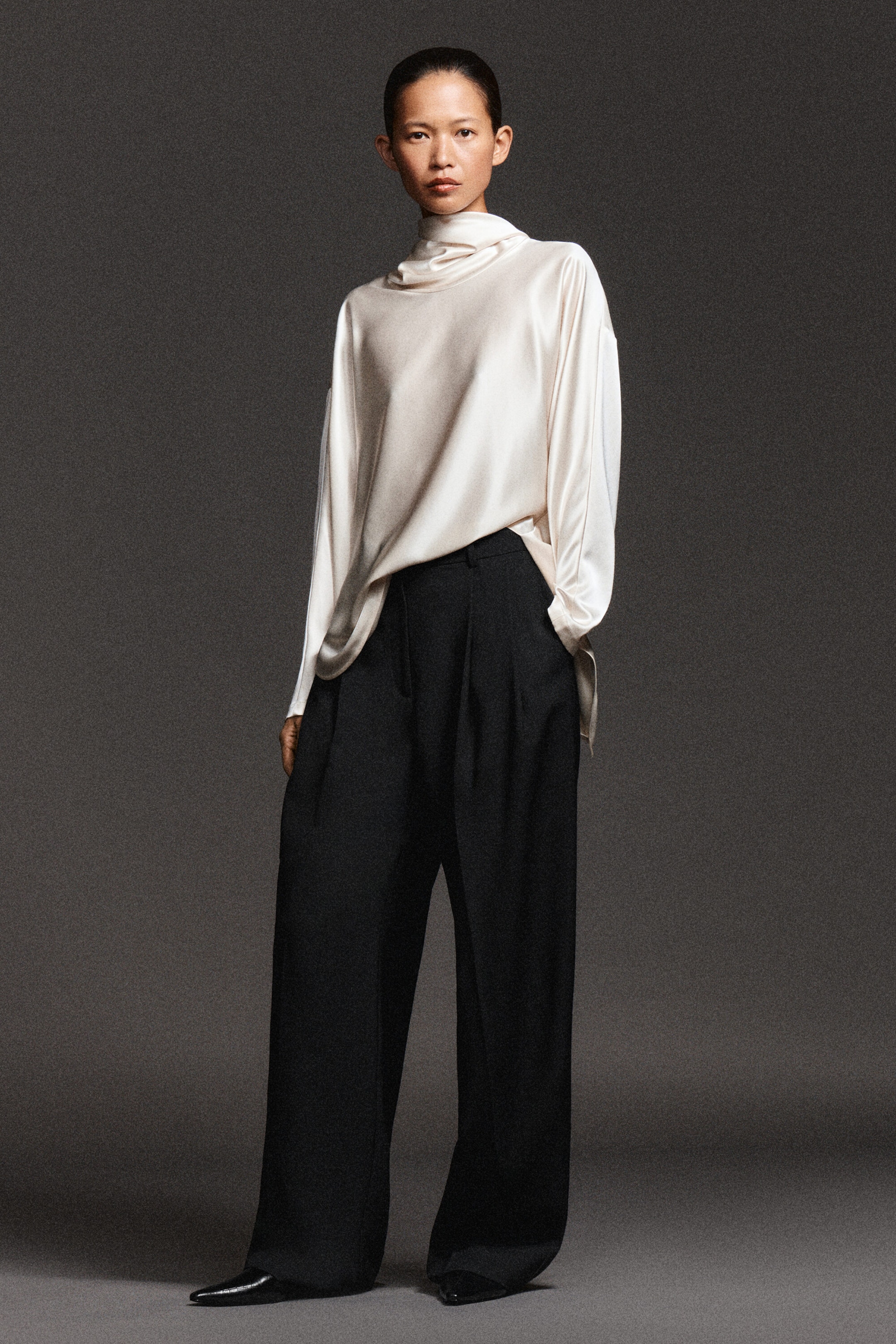 A cream satin blouse featuring a draped cowl neck and long, loose sleeves is styled partially tucked into high-waisted, wide-leg black trousers with pleated details. Facing front, the outfit presents a relaxed silhouette, highlighting the fluid drape of the satin fabric against the structured design of the trousers. A cream satin blouse featuring a draped cowl neck and long, loose sleeves is styled partially tucked into high-waisted, wide-leg black trousers with pleated details. Facing front, the outfit presents a relaxed silhouette, highlighting the fluid drape of the satin fabric against the structured design of the trousers.