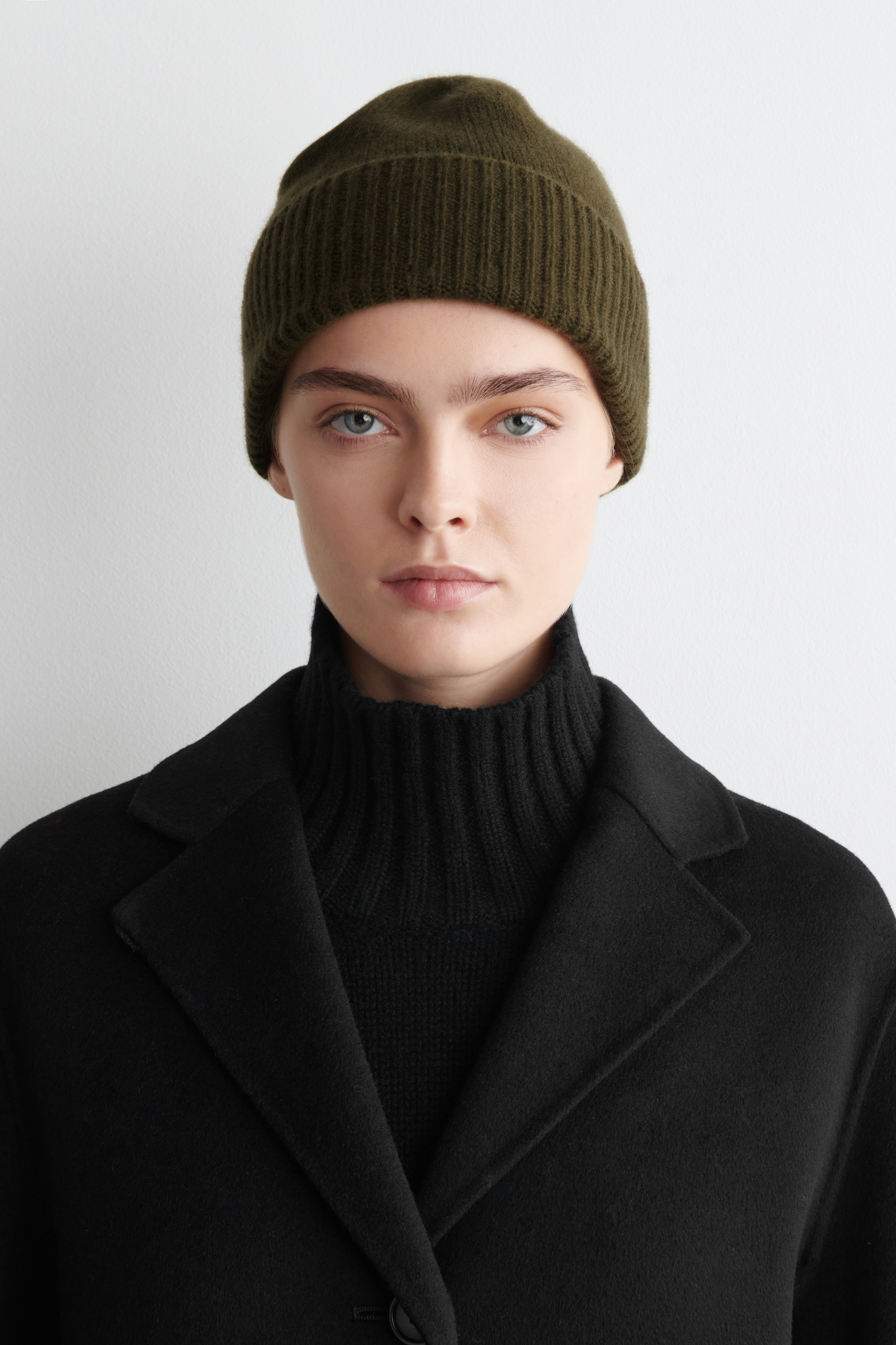 Größeres Bild ansehen: Eine olivgrüne Strickmütze wird frontseitig getragen, mit einem gerippten, umgeschlagenen Rand und kombiniert mit einem schwarzen gerippten Rollkragenpullover und Mantel.