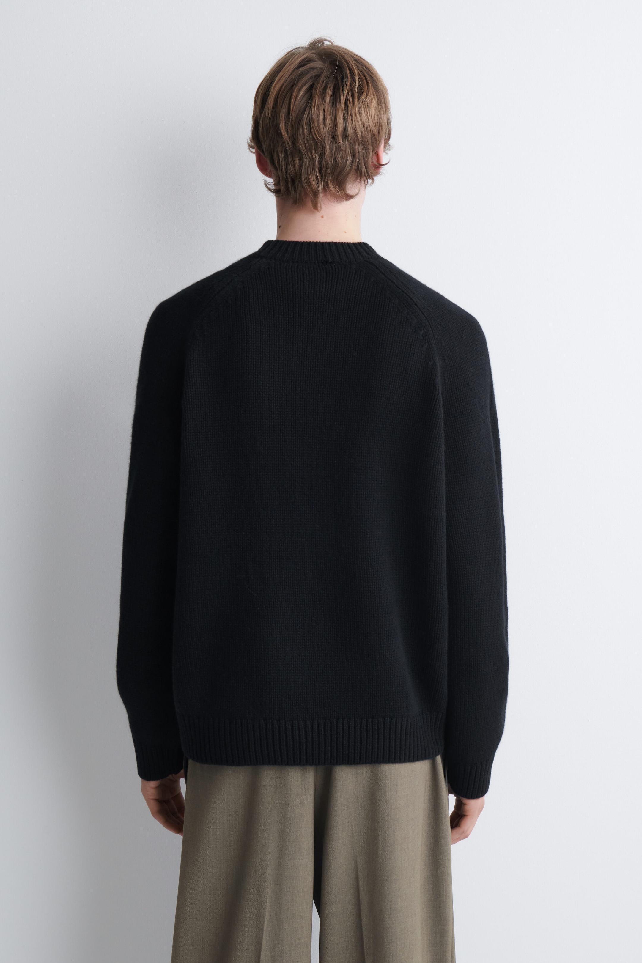 A black knit jumper featuring a crew neck, raglan sleeves and a ribbed hem is being worn by the model, shown from the back. The jumper is styled with olive-green trousers. A black knit jumper featuring a crew neck, raglan sleeves and a ribbed hem is being worn by the model, shown from the back. The jumper is styled with olive-green trousers.