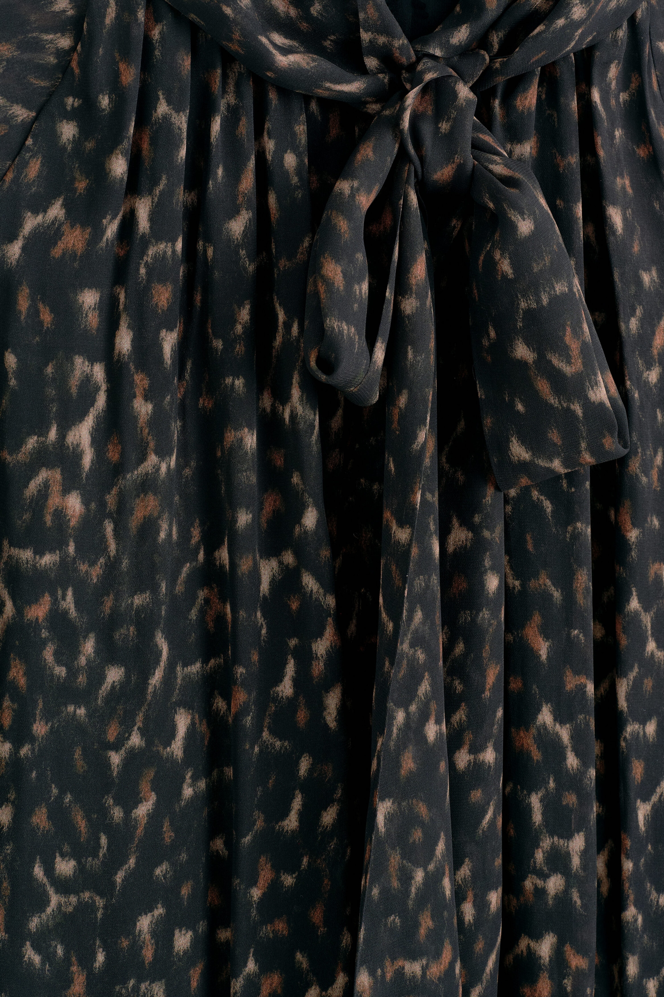 The leopard-print fluid scarf-detail dress features a dark, abstract animal print in black, brown, and beige tones. A matching fabric scarf detail is tied in a loose knot at the centre, with the fluid, lightweight fabric draping downward in soft, gathered folds. The leopard-print fluid scarf-detail dress features a dark, abstract animal print in black, brown, and beige tones. A matching fabric scarf detail is tied in a loose knot at the centre, with the fluid, lightweight fabric draping downward in soft, gathered folds.