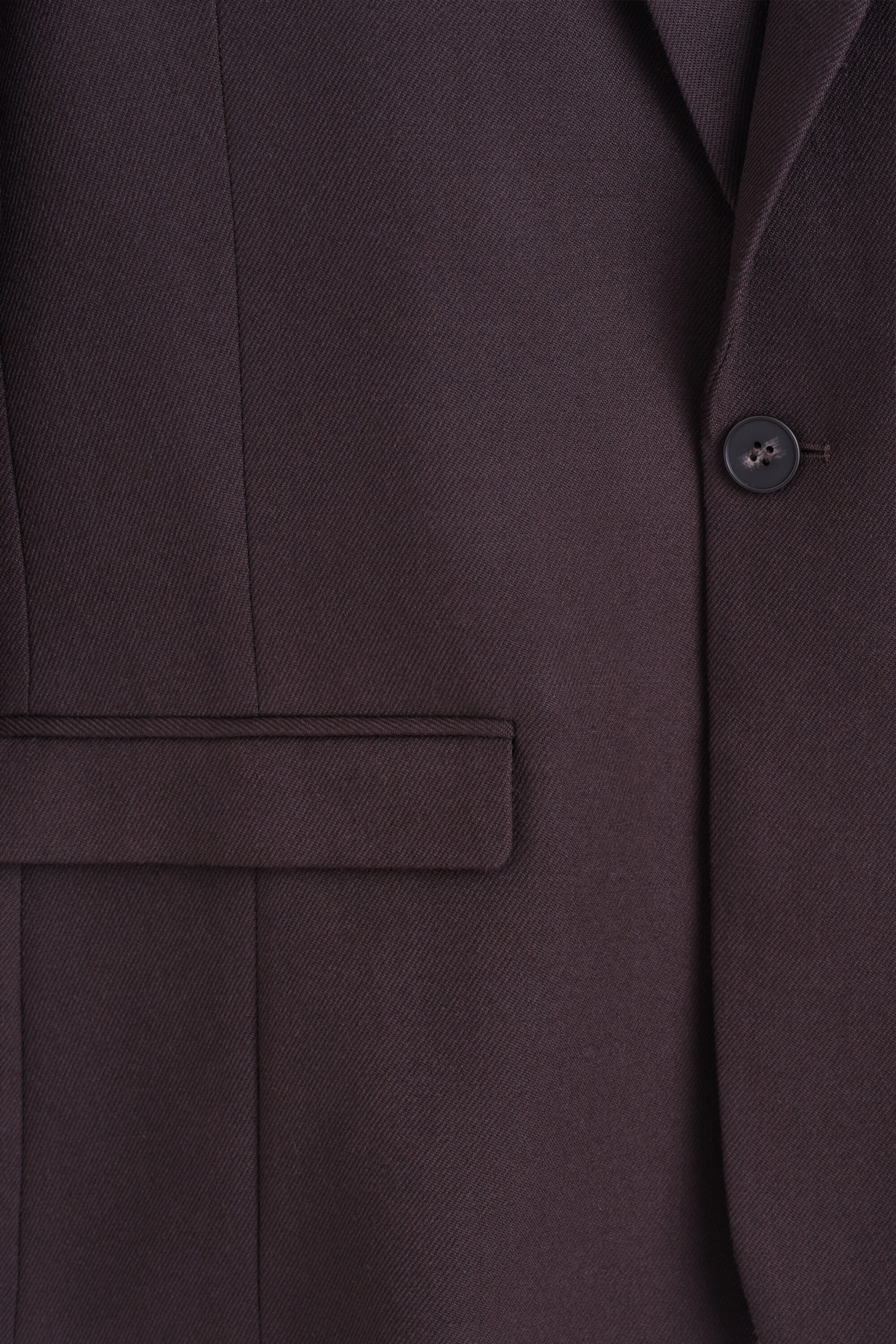 A dark brown suit jacket is shown from the front, featuring a subtle diagonal weave, a single dark button fastening on the right, and a flap pocket on the left. A dark brown suit jacket is shown from the front, featuring a subtle diagonal weave, a single dark button fastening on the right, and a flap pocket on the left.