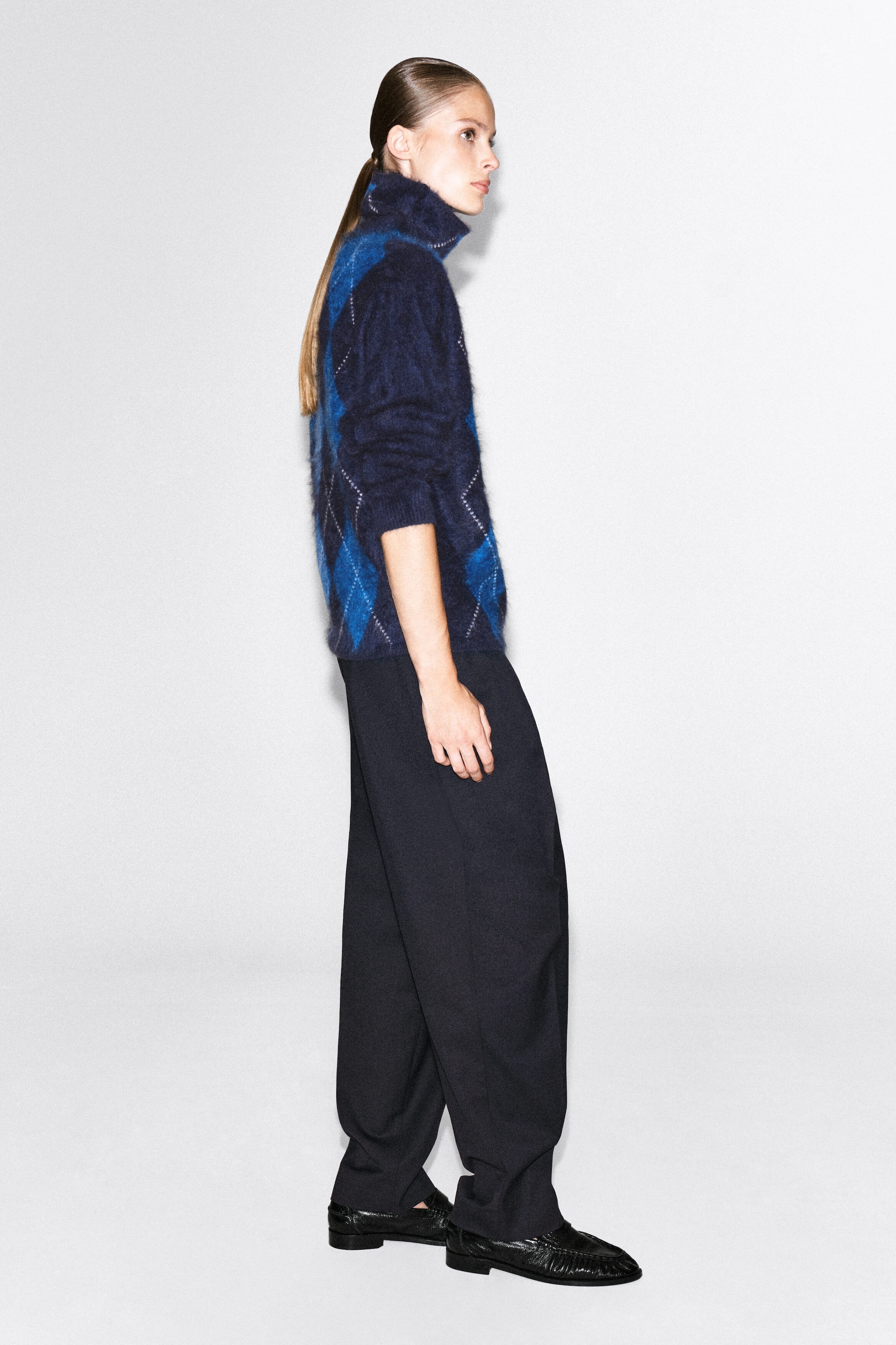 A brushed pure cashmere argyle funnel-neck jumper in shades of navy and bright blue is worn by the model. The fuzzy, textured knitwear is paired with dark, loose-fitting trousers and black flat shoes. A brushed pure cashmere argyle funnel-neck jumper in shades of navy and bright blue is worn by the model. The fuzzy, textured knitwear is paired with dark, loose-fitting trousers and black flat shoes.