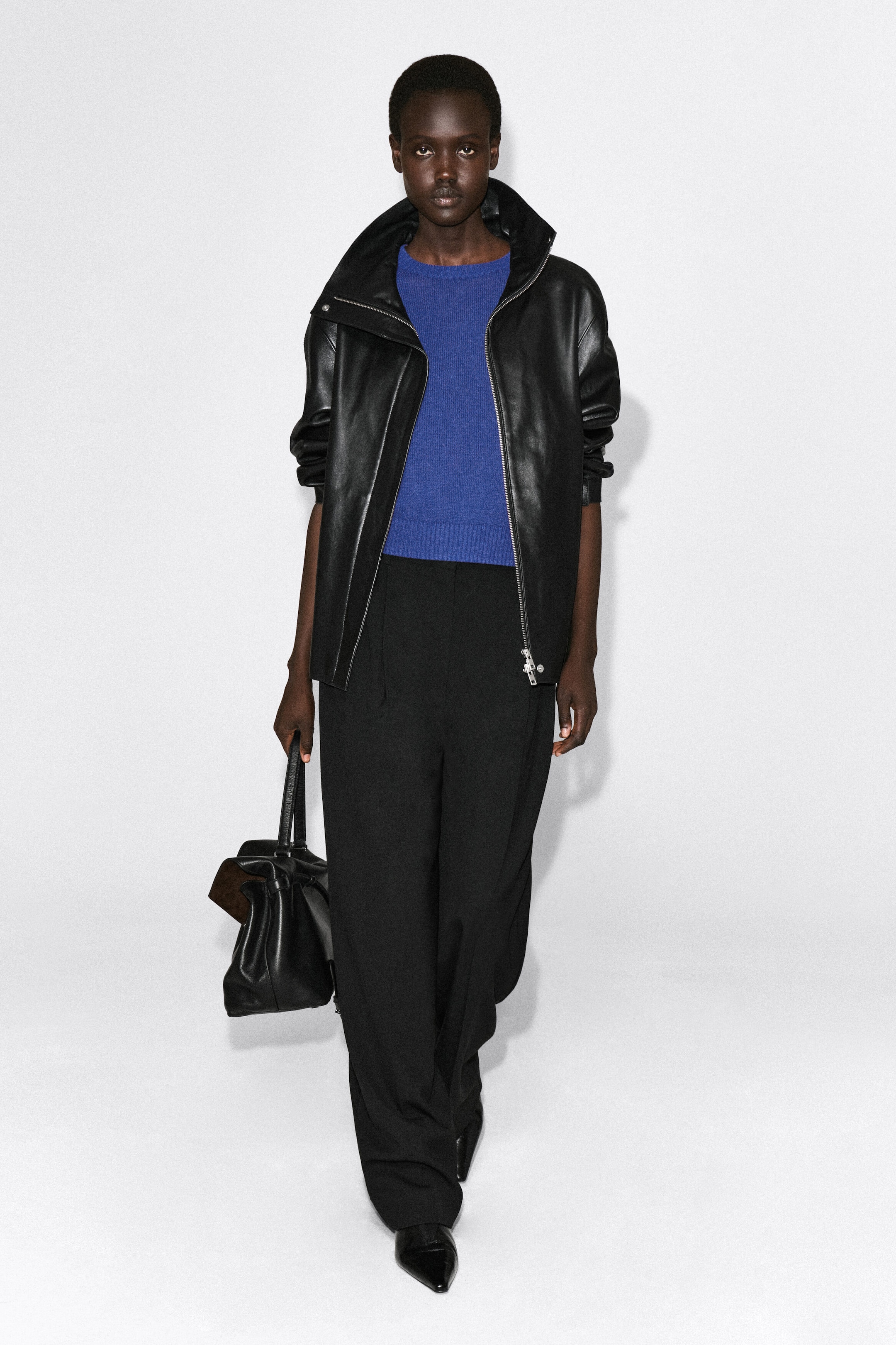 The shrunken knitted linen T-shirt in a vibrant blue is shown from the front, featuring a fine knit texture, a crew neck, and a ribbed hem. It fits closely in a cropped length, layered underneath an open black leather jacket to highlight its structured shape.