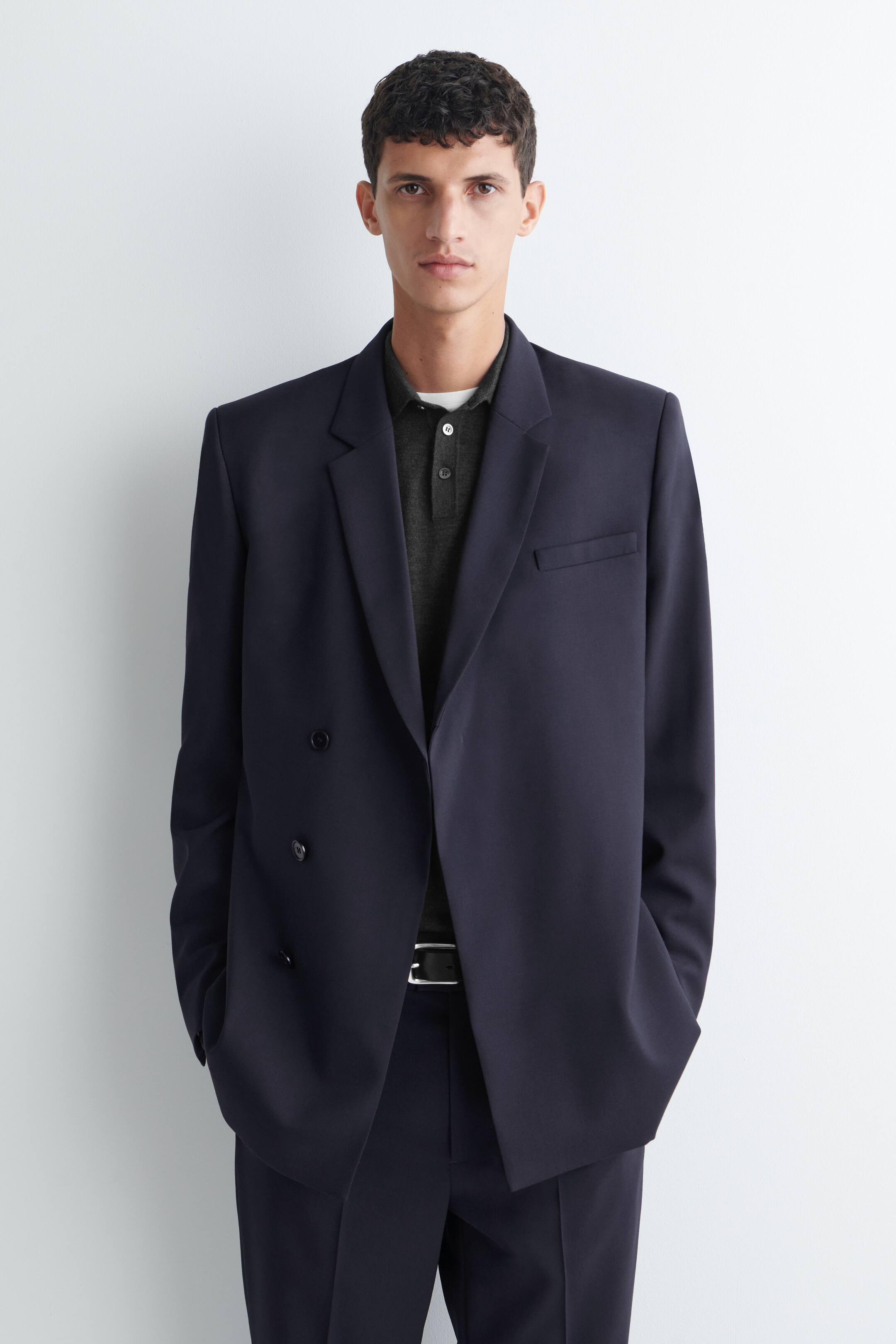 Agrandir l'image: Un blazer en laine bleu marine à double boutonnage, avec des revers crantés, une poche poitrine et des boutons décoratifs sur le devant, est porté par le mannequin. Le blazer est superposé à un polo gris foncé et associé à un pantalon bleu marine assorti.