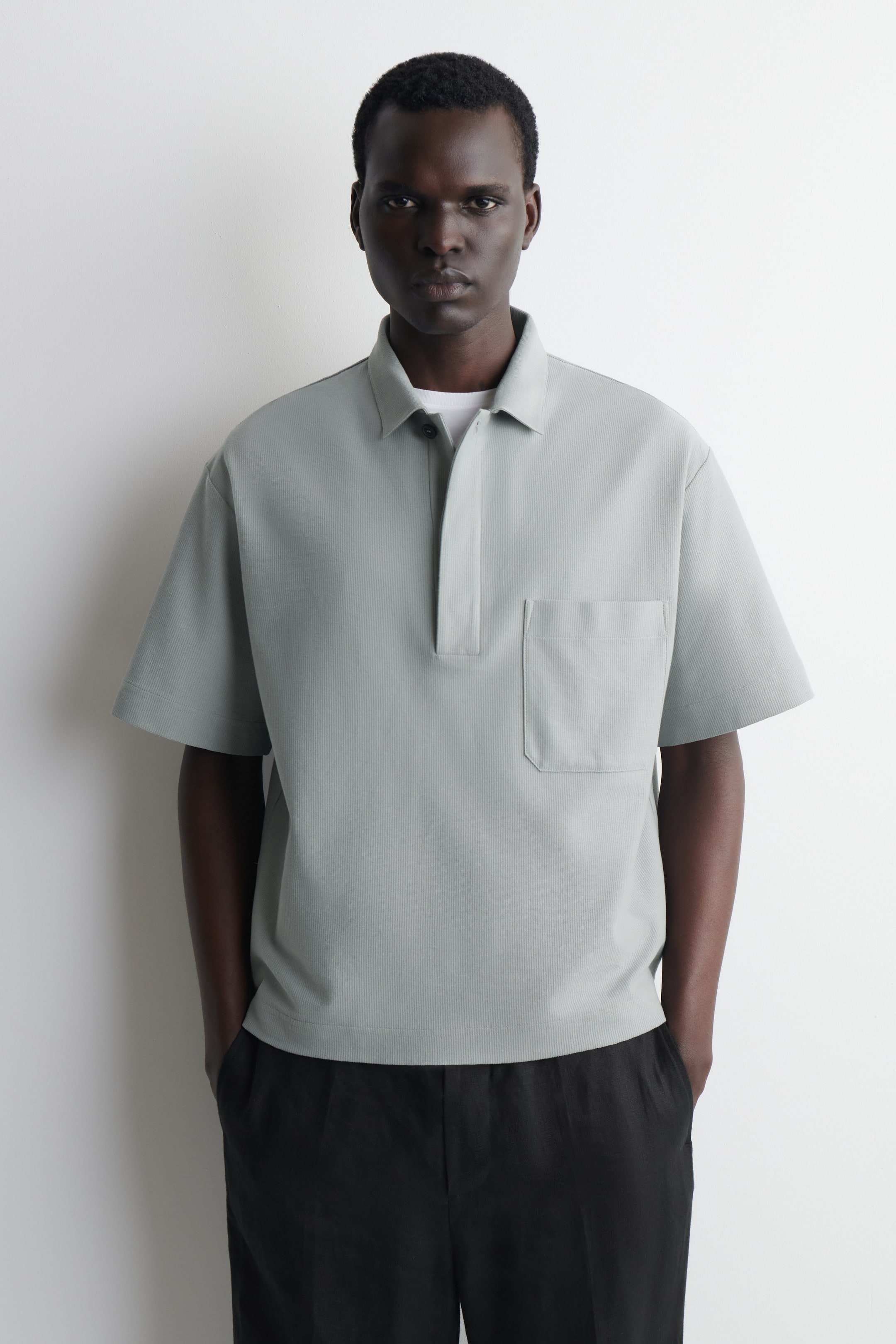 Ver imagen más grande: Se presenta una camiseta polo de color verde claro vista desde el frente, con un cuello abierto, una tapeta corta de dos botones con un botón oscuro visible, y mangas cortas anchas. Debajo del cuello se aprecia una camiseta interior blanca, y la camisa muestra un bolsillo de parche en el pecho izquierdo con un sutil patrón texturizado vertical a lo largo de su tejido.