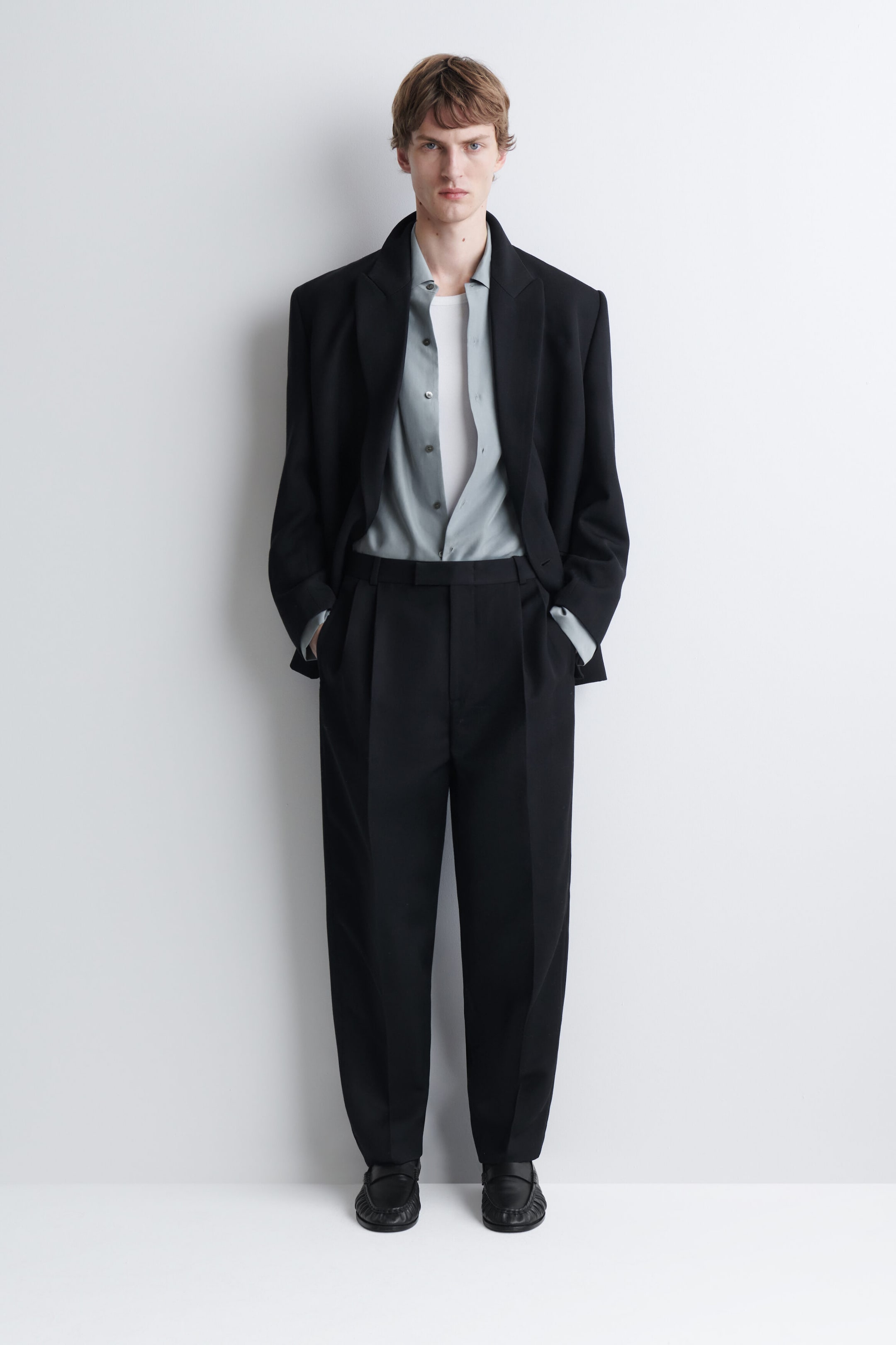 The model is wearing a relaxed silk shirt in a light grey shade, tucked into black trousers and layered under an open black blazer. The shirt is styled unbuttoned at the collar to reveal a white undershirt. The model is wearing a relaxed silk shirt in a light grey shade, tucked into black trousers and layered under an open black blazer. The shirt is styled unbuttoned at the collar to reveal a white undershirt.