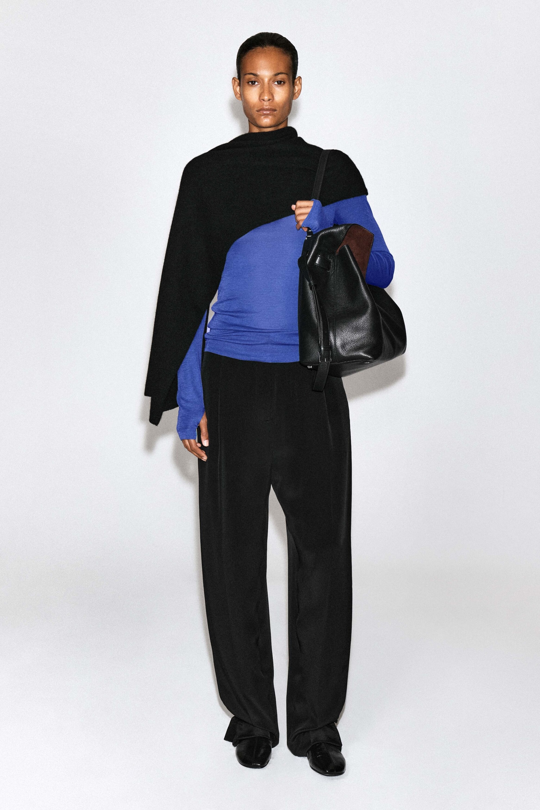 A slim-fitting, long-sleeved blue merino wool top is worn by the model, layered underneath an asymmetrical black knit wrap draped over one shoulder. The top is styled with wide-leg black trousers, and the model is carrying a large black leather shoulder bag. A slim-fitting, long-sleeved blue merino wool top is worn by the model, layered underneath an asymmetrical black knit wrap draped over one shoulder. The top is styled with wide-leg black trousers, and the model is carrying a large black leather shoulder bag.