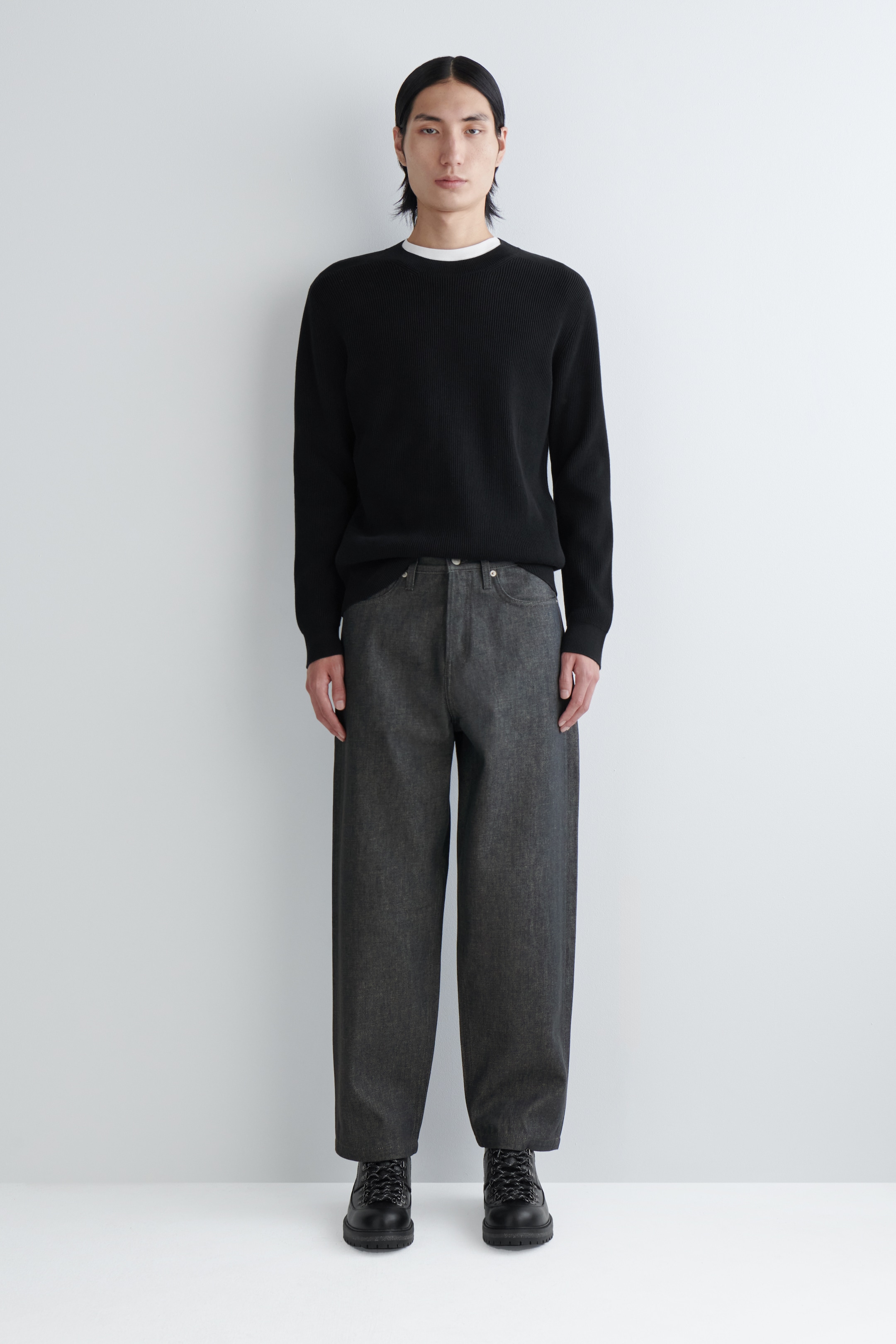 Agrandir l'image: Un pull noir à col rond côtelé est porté par le mannequin, superposé à un t-shirt blanc dont le col est visible au niveau de l'encolure. Les poignets et l'ourlet côtelés du pull sont visibles, et il est assorti à un pantalon gris foncé.