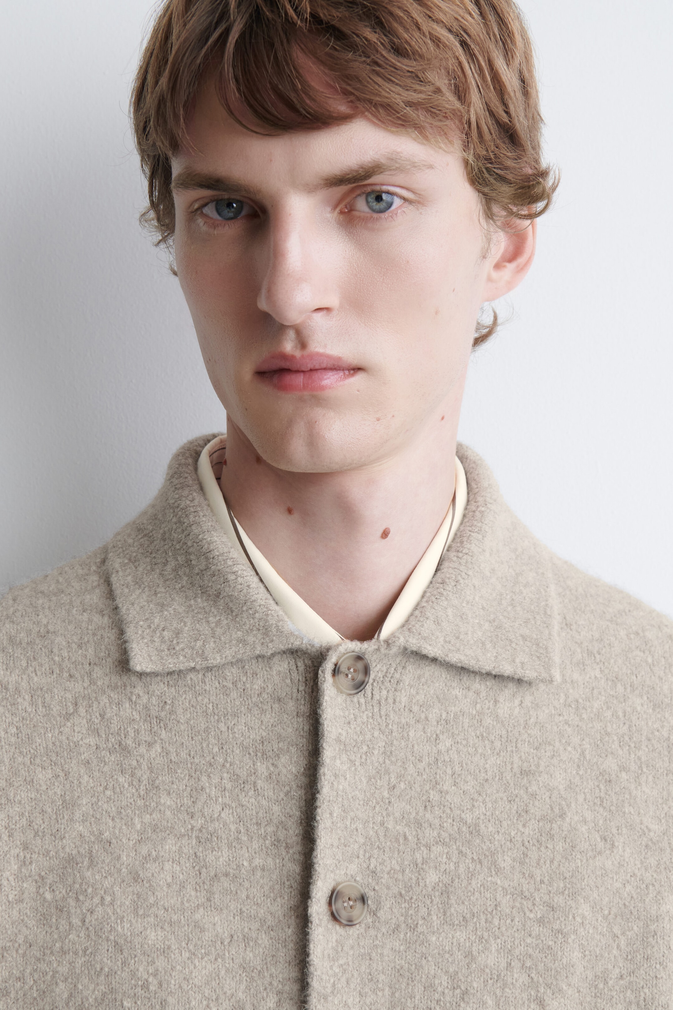 A light beige brushed-knit cardigan with a classic collar and front buttons is being worn by the model. The cardigan is layered over a cream-coloured collared shirt. A light beige brushed-knit cardigan with a classic collar and front buttons is being worn by the model. The cardigan is layered over a cream-coloured collared shirt.