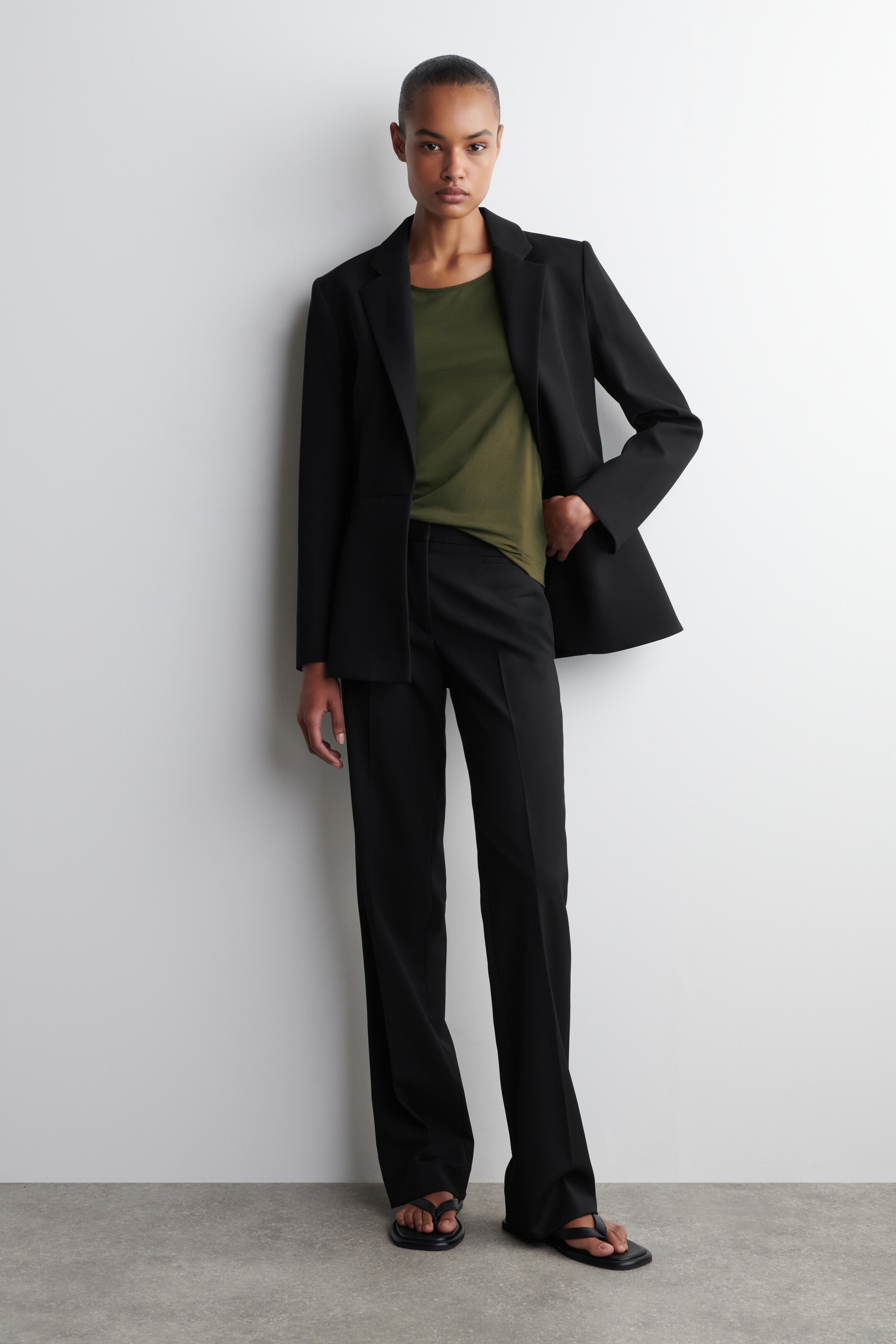 Größeres Bild anzeigen: Ein schwarzer einreihiger Blazer wird offen getragen, wodurch ein olivgrünes Rundhals-Top sichtbar wird, das teilweise in schwarze maßgeschneiderte Hosen gesteckt ist. Der Blazer hat Kerbrevers und eine lockere Passform, und die Hosen zeigen einen geraden Schnitt mit einer sichtbaren vorderen Bügelfalte, alles kombiniert mit schwarzen flachen Sandalen.