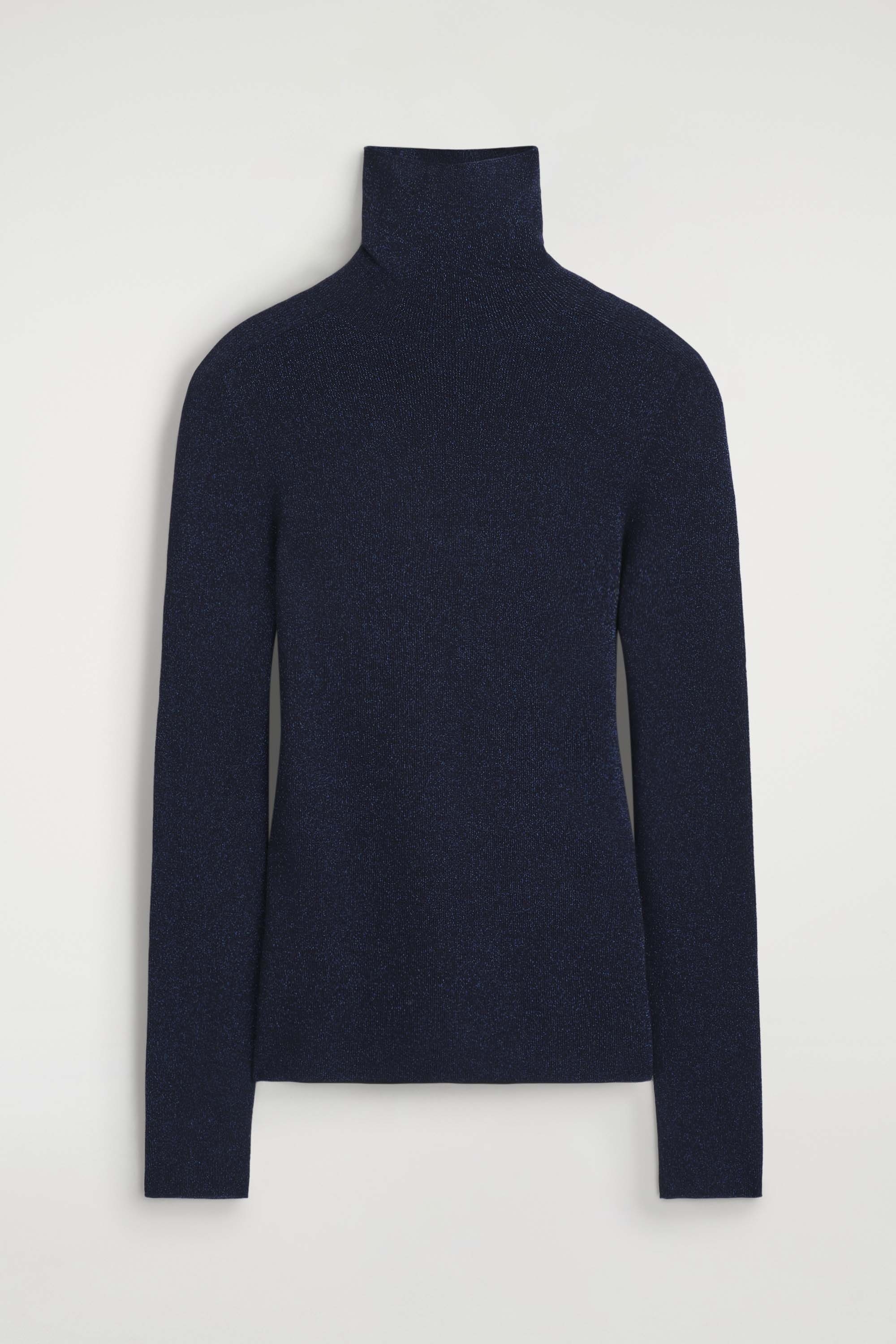 A model is showcasing the sparkly seamless wool funnel-neck top from the front, highlighting its slim-fitting silhouette and shimmering dark blue knit fabric. The top features a high, folded funnel neck and long sleeves, styled untucked over black trousers.