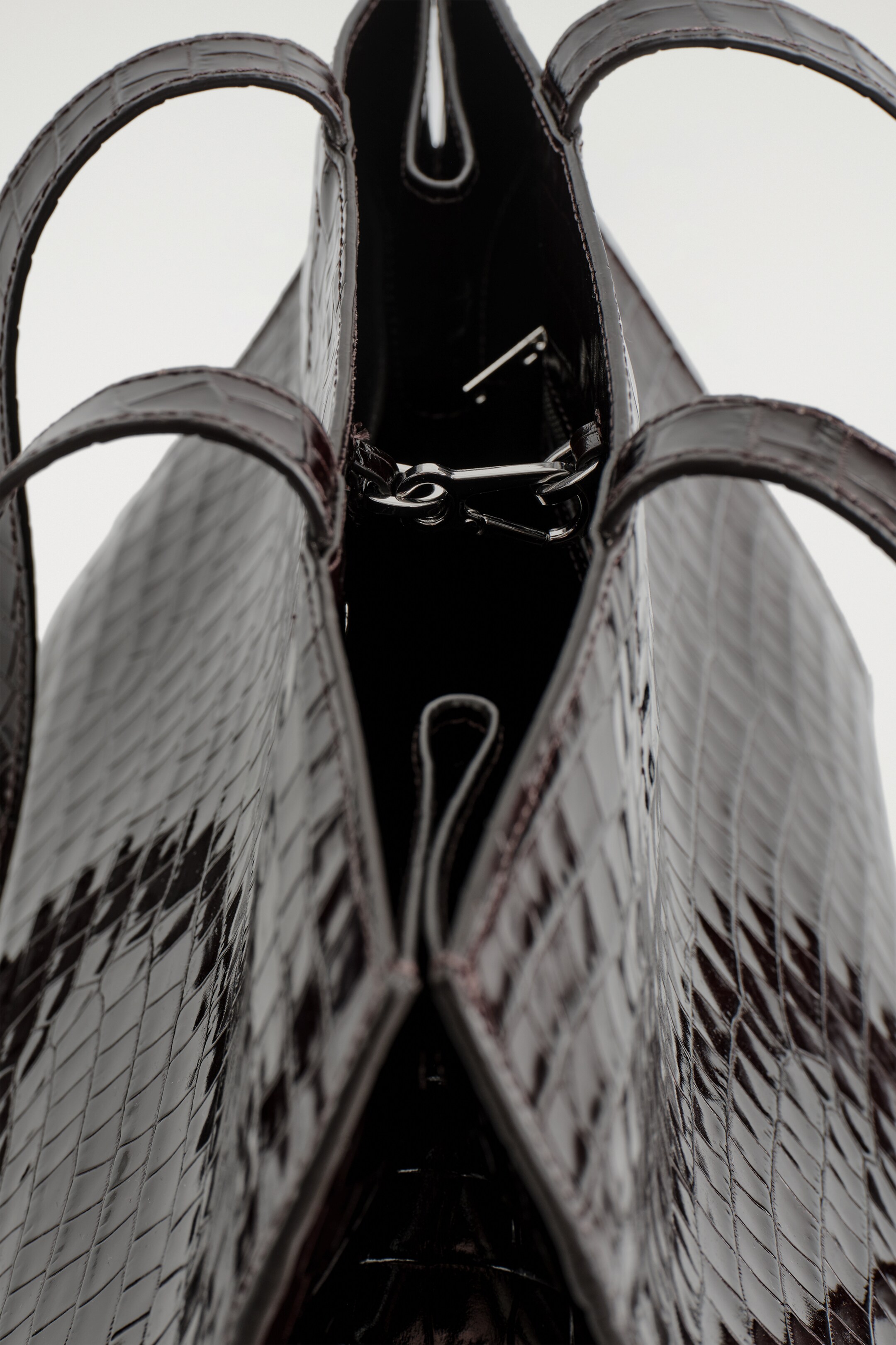 Agrandir l'image: Un sac fourre-tout en cuir embossé façon croco, sombre et brillant, est vu depuis le HAUT, avec son ouverture tournée vers le haut, doté de longues bretelles et d'une forme pincée grâce à des plis subtils sur les côtés. Une fermeture à clip de couleur argentée et une poche intérieure sont visibles à l'intérieur sombre.