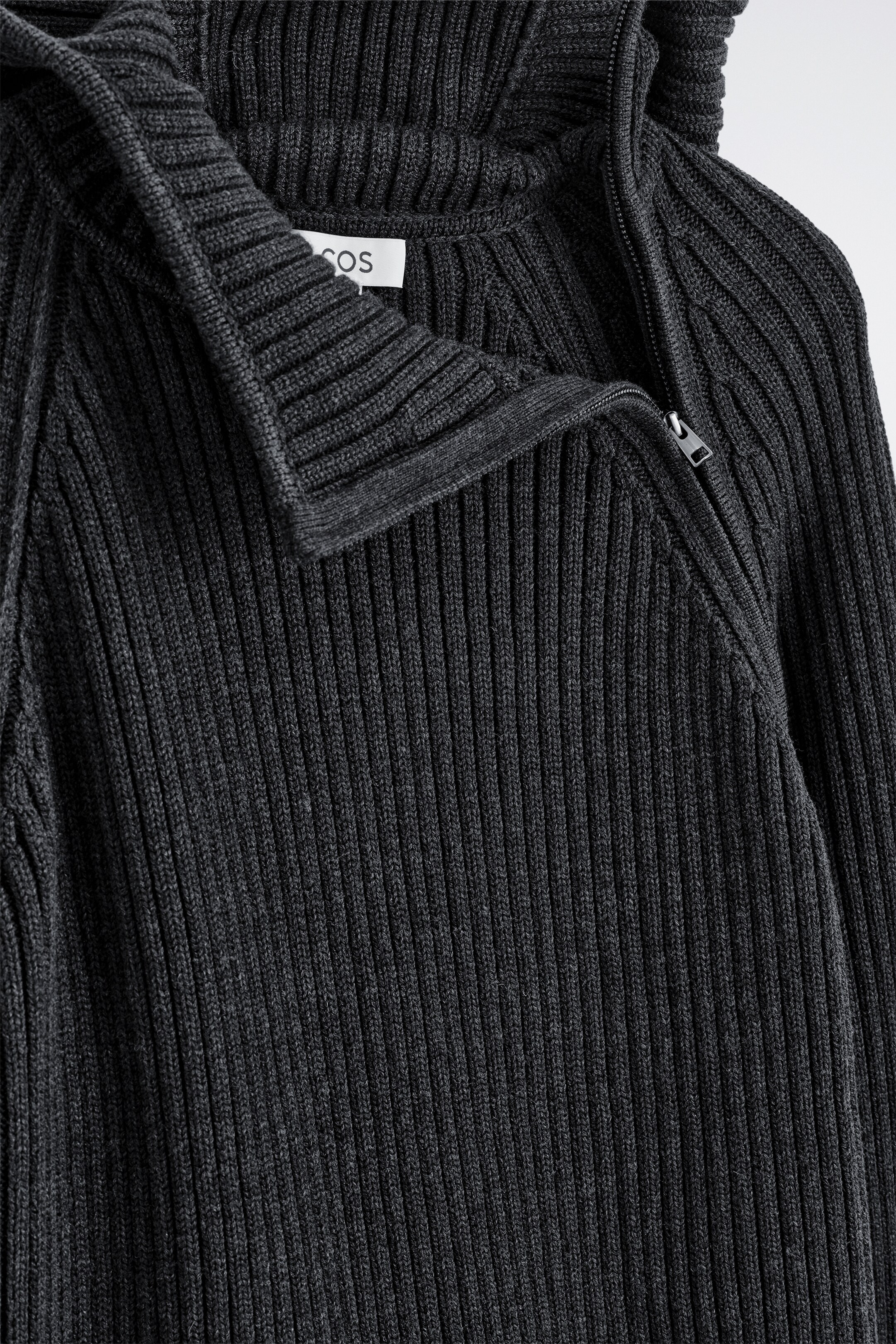Agrandir l'image: Un hoodie mélange de charbon est présenté, tricoté dans une confection à larges côtes. Il est doté d'une capuche tubulaire avec une fermeture à glissière partielle qui court en diagonale depuis l'épaule droite jusqu'à la partie supérieure de la poitrine. Une étiquette blanche COS est visible à l'intérieur du col.