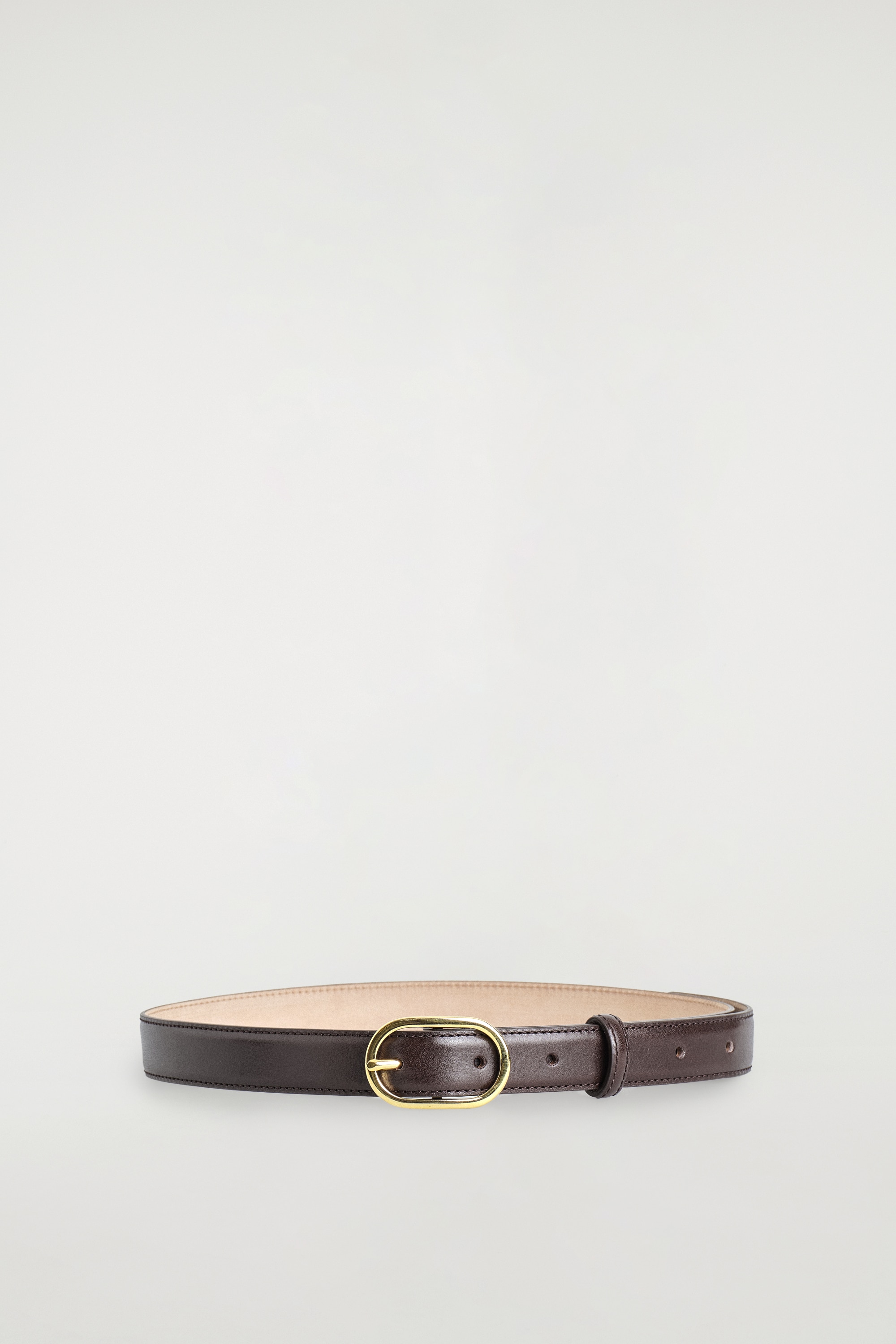 Een donkerbruine leren riem met een gepolijste ovale gesp in goudtint wordt door het model gedragen door de lussen van een lichtgewassen denim jeans. De riem wordt getoond onder een gedeeltelijk ingestopte witte gebreide trui.