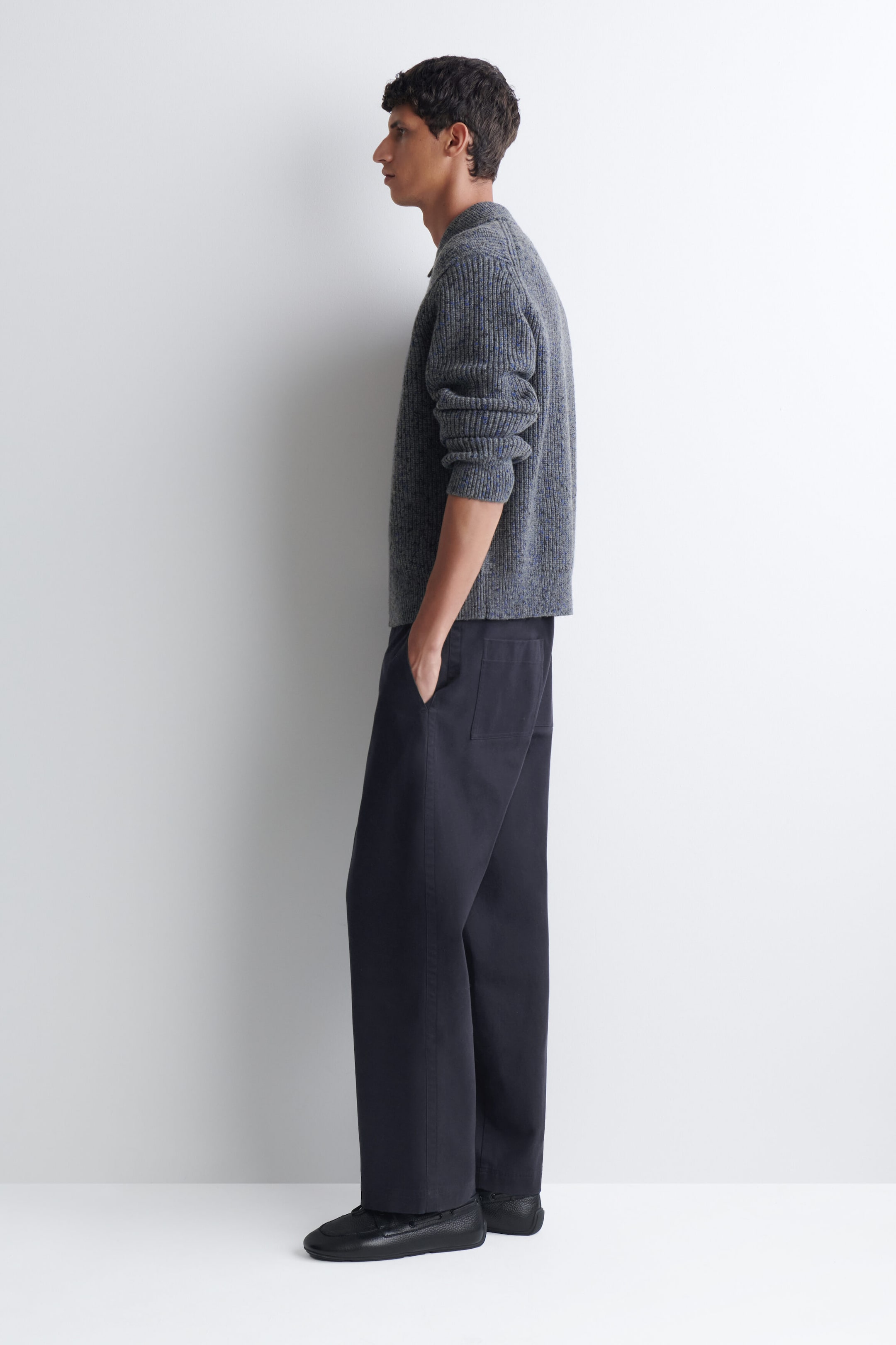 A grey, ribbed-knit polo sweater with pushed-up sleeves is worn by the model, styled with dark, wide-leg trousers. The model is standing in profile with one hand resting in the trouser pocket. A grey, ribbed-knit polo sweater with pushed-up sleeves is worn by the model, styled with dark, wide-leg trousers. The model is standing in profile with one hand resting in the trouser pocket.