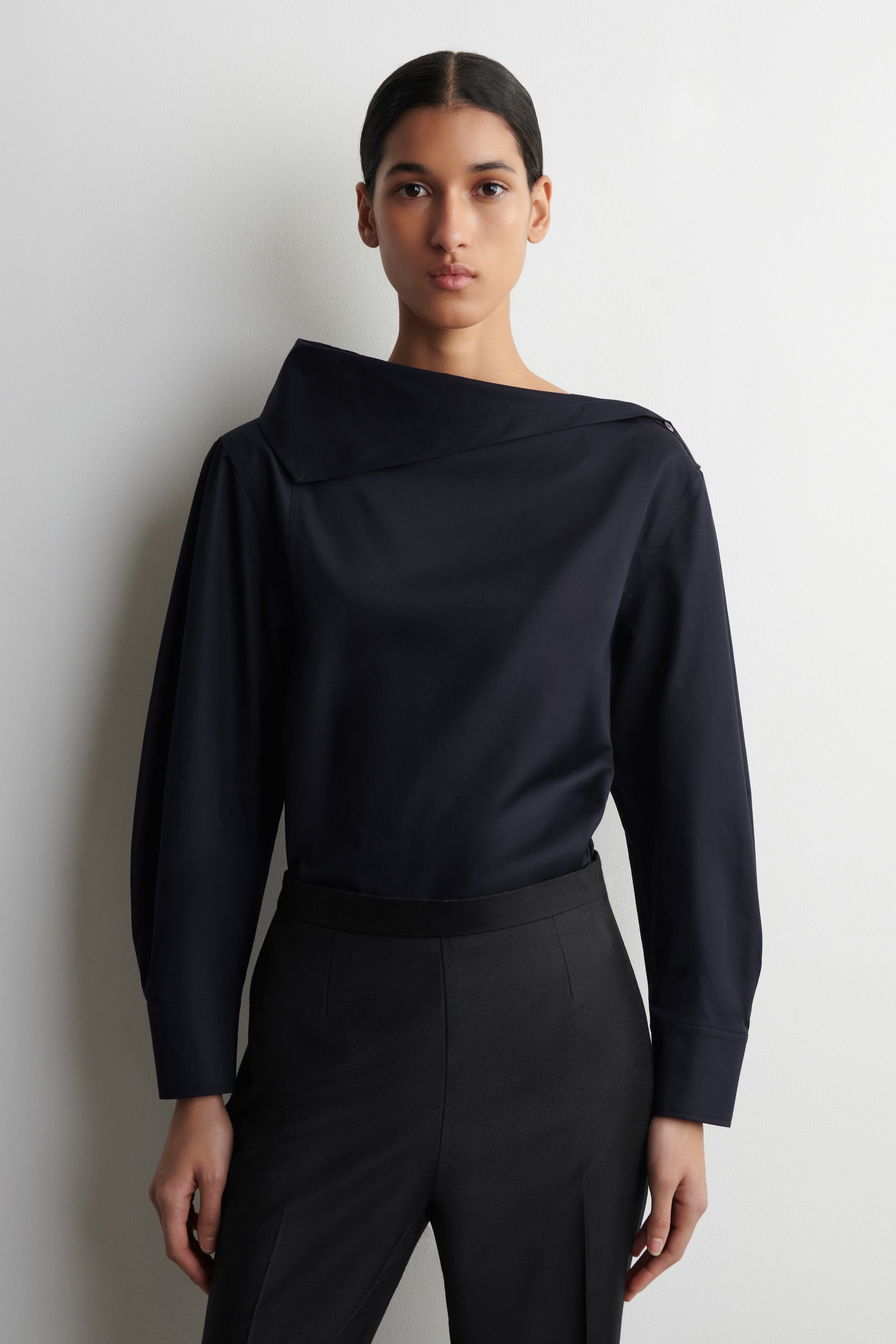 הצגת תמונה גדולה יותר: חולצה כחולה כהה מציגה עיצוב אסימטרי עם צווארון רחב ומובנה המתקפל מעל הכתף השמאלית ומחשוף פתוח בצד ימין. היא כוללת שרוולים ארוכים עם חפתים מכופתרים ונלבשת בתוך מכנסיים שחורים, נראית מקדימה, בזווית קלה ימינה.