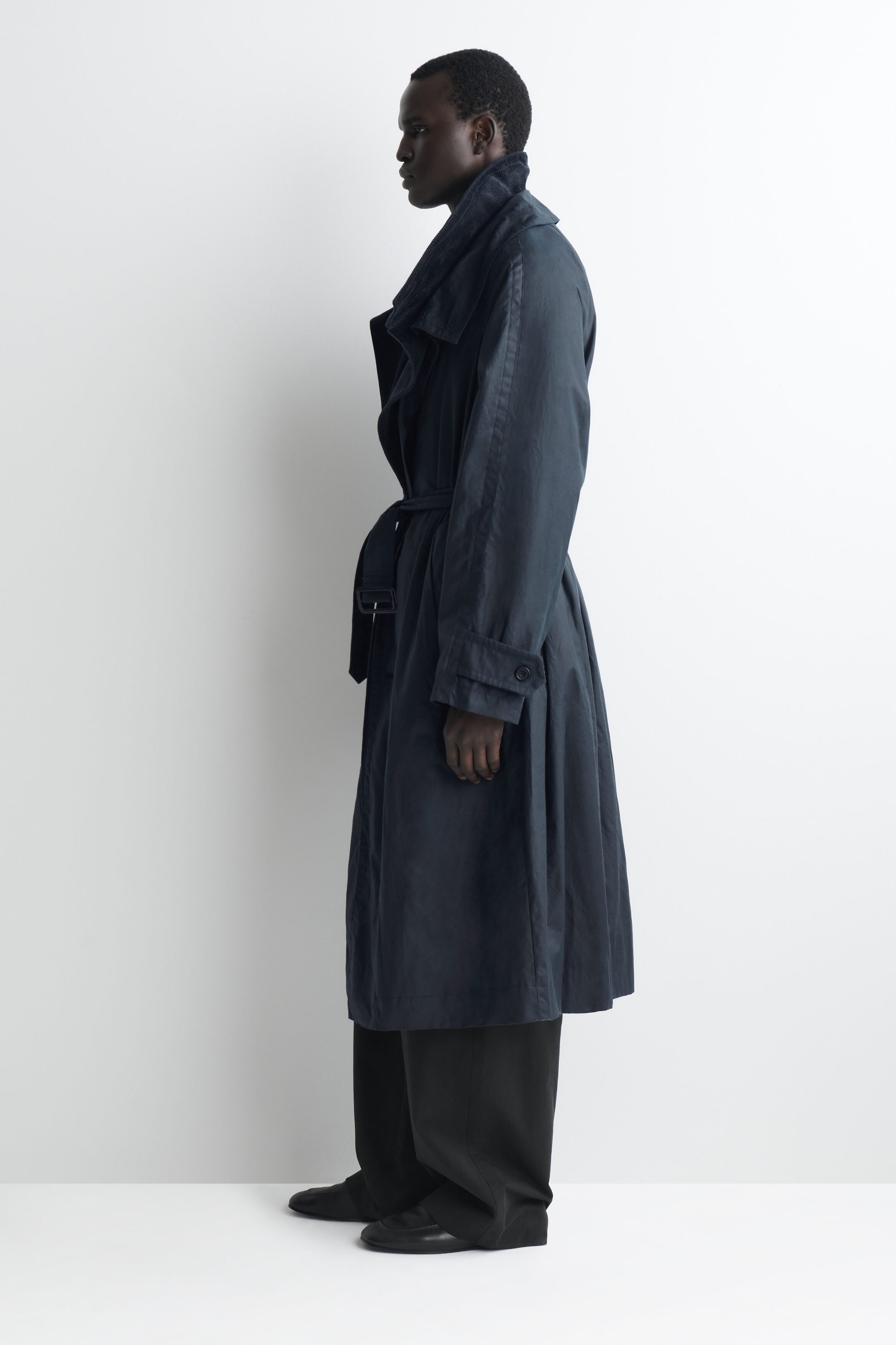 A long, midnight-blue trench coat is shown in left-facing profile, highlighting its relaxed fit and midi length. The coat features a belted waist, buttoned cuff tabs, and a high funnel-neck collar with tonal corduroy lining, styled over wide-leg black trousers. A long, midnight-blue trench coat is shown in left-facing profile, highlighting its relaxed fit and midi length. The coat features a belted waist, buttoned cuff tabs, and a high funnel-neck collar with tonal corduroy lining, styled over wide-leg black trousers.