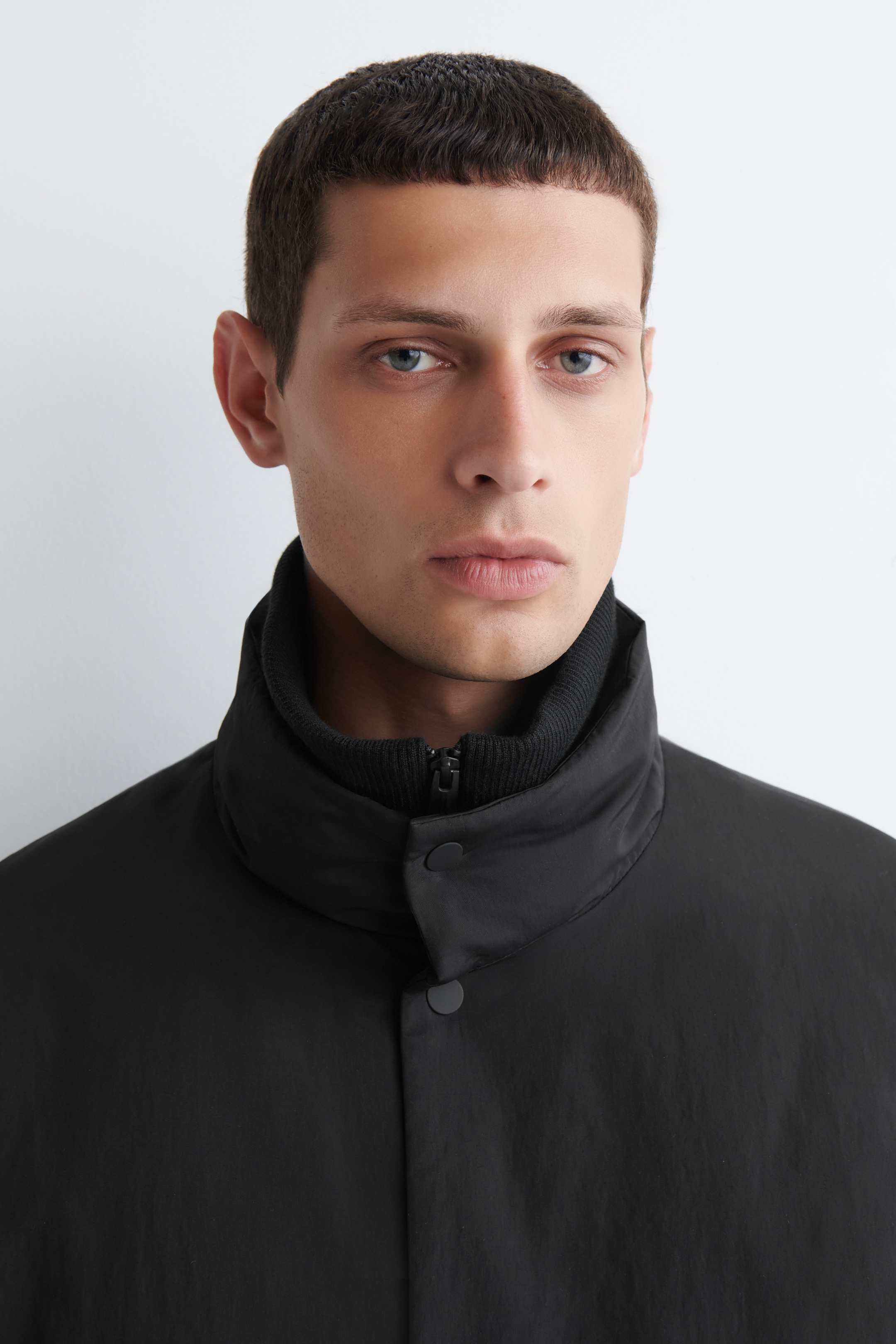 Größeres Bild ansehen: Eine schwarze Jacke wird gezeigt, die einen markanten Stehkragen und zwei sichtbare Druckknopfverschlüsse an der Vorderseite aufweist. Ein schwarzer gerippter Strickkragen ist darunter sichtbar, mit seinem Reißverschluss, der am Hals zu sehen ist.