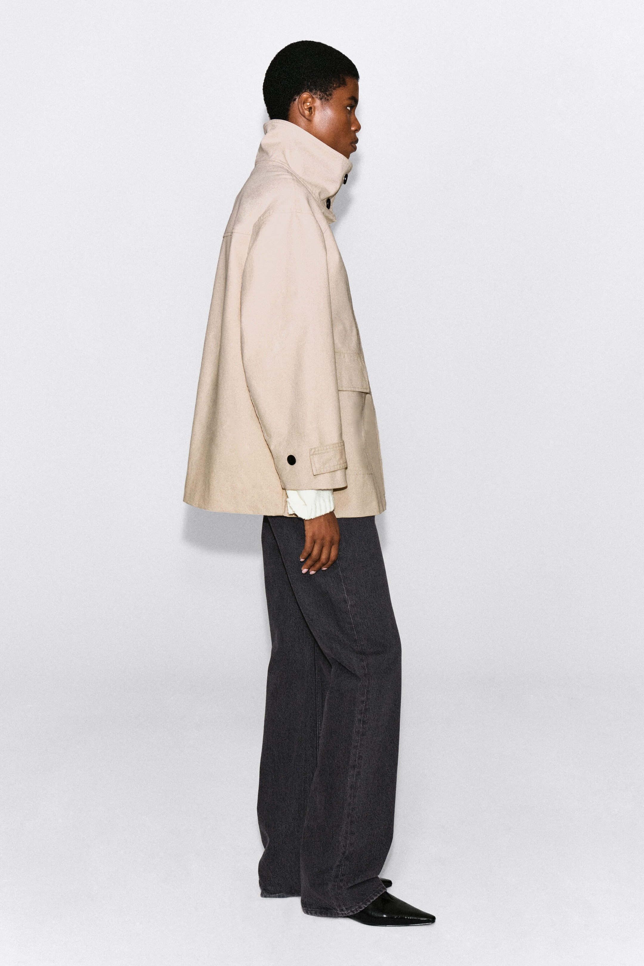 A beige cotton twill funnel-neck jacket is worn by the model, highlighting its high structured collar and relaxed A-line silhouette in side profile. The jacket features button-tab cuffs layered over a white knit sleeve and is styled with dark wide-leg trousers. A beige cotton twill funnel-neck jacket is worn by the model, highlighting its high structured collar and relaxed A-line silhouette in side profile. The jacket features button-tab cuffs layered over a white knit sleeve and is styled with dark wide-leg trousers.