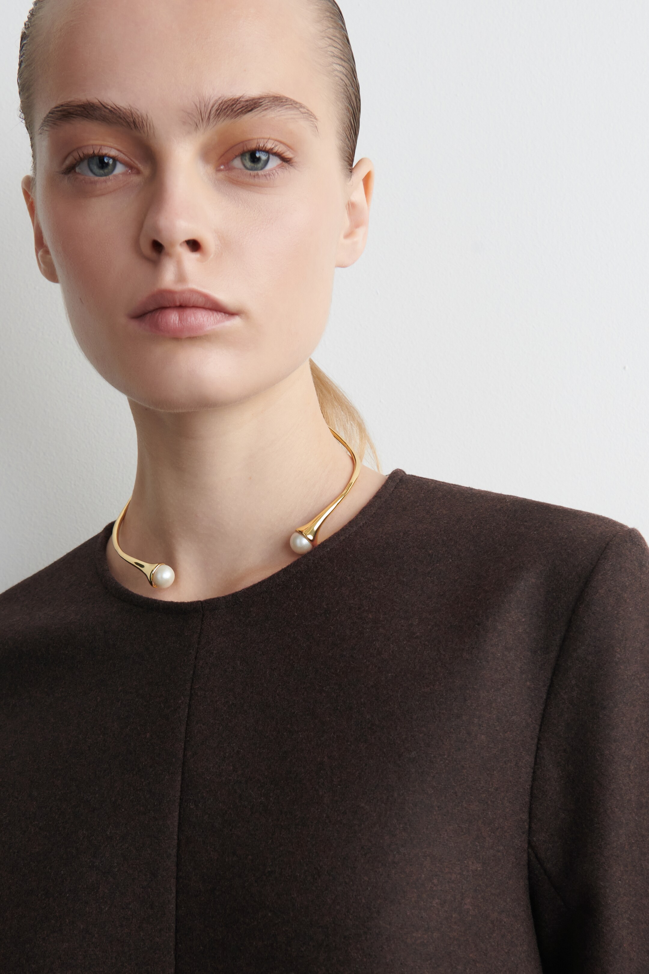 Ver imagen más grande: Un collar de tono dorado se lleva alrededor del cuello, con sus dos extremos ensanchados, cada uno con una perla blanca, colocados en la parte delantera. El collar descansa sobre una prenda marrón oscuro.