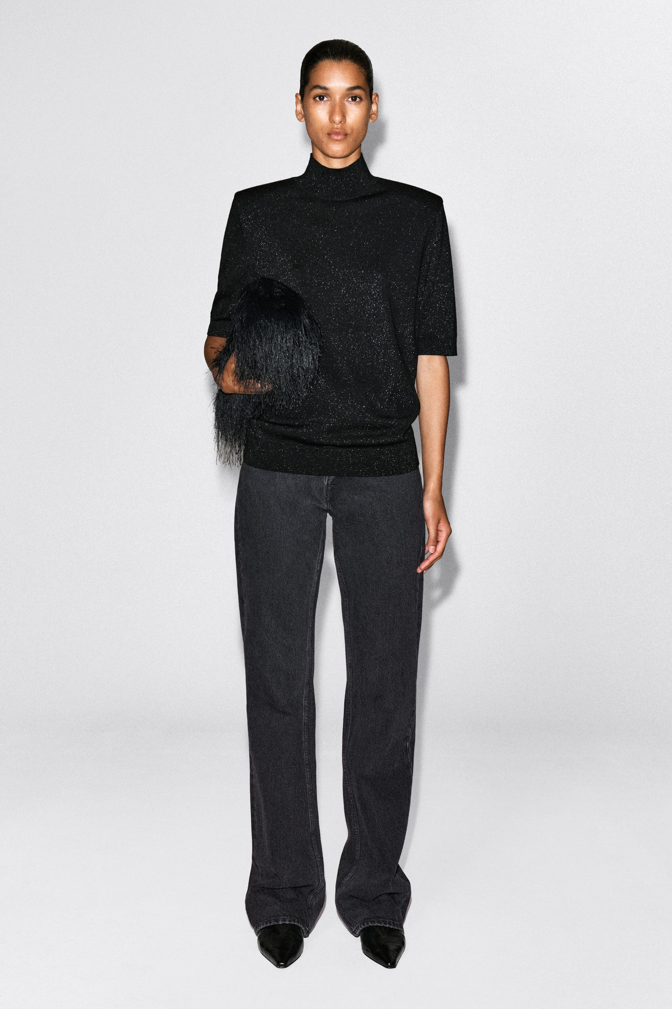 A black, fine-knit mock-neck top featuring shimmering metallic flecks is shown from the front, showcasing structured, sculpted shoulders and elbow-length sleeves. The top is fitted with a ribbed hem and is styled with dark grey jeans. A black, fine-knit mock-neck top featuring shimmering metallic flecks is shown from the front, showcasing structured, sculpted shoulders and elbow-length sleeves. The top is fitted with a ribbed hem and is styled with dark grey jeans.