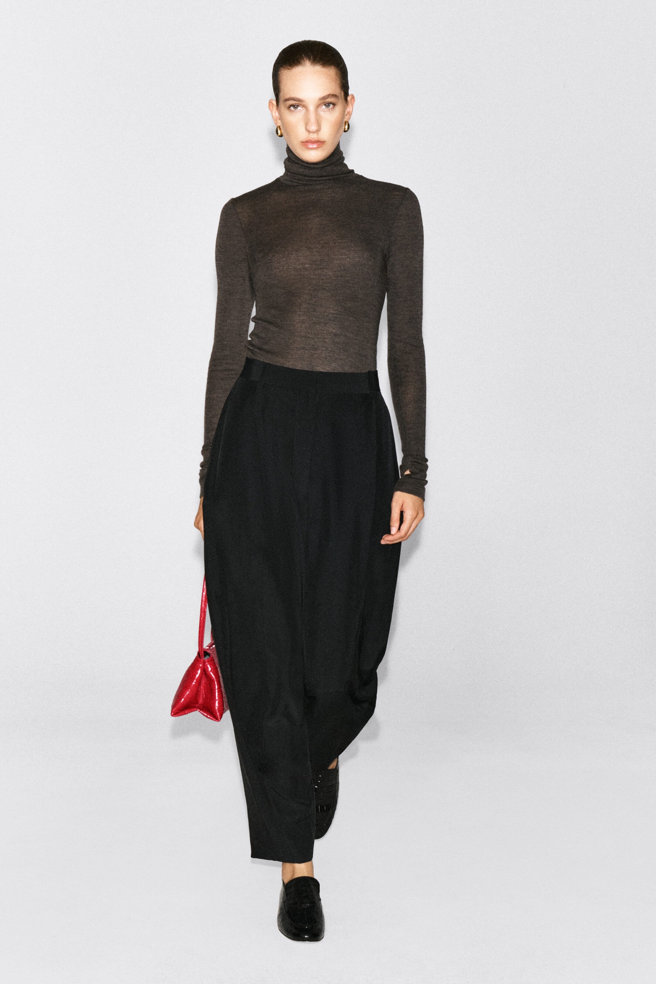 A dark brown merino wool high-neck top with a fine-knit, semi-sheer texture is worn by the model. The long-sleeved turtleneck top is tucked into a pair of high-waisted black trousers. A dark brown merino wool high-neck top with a fine-knit, semi-sheer texture is worn by the model. The long-sleeved turtleneck top is tucked into a pair of high-waisted black trousers.