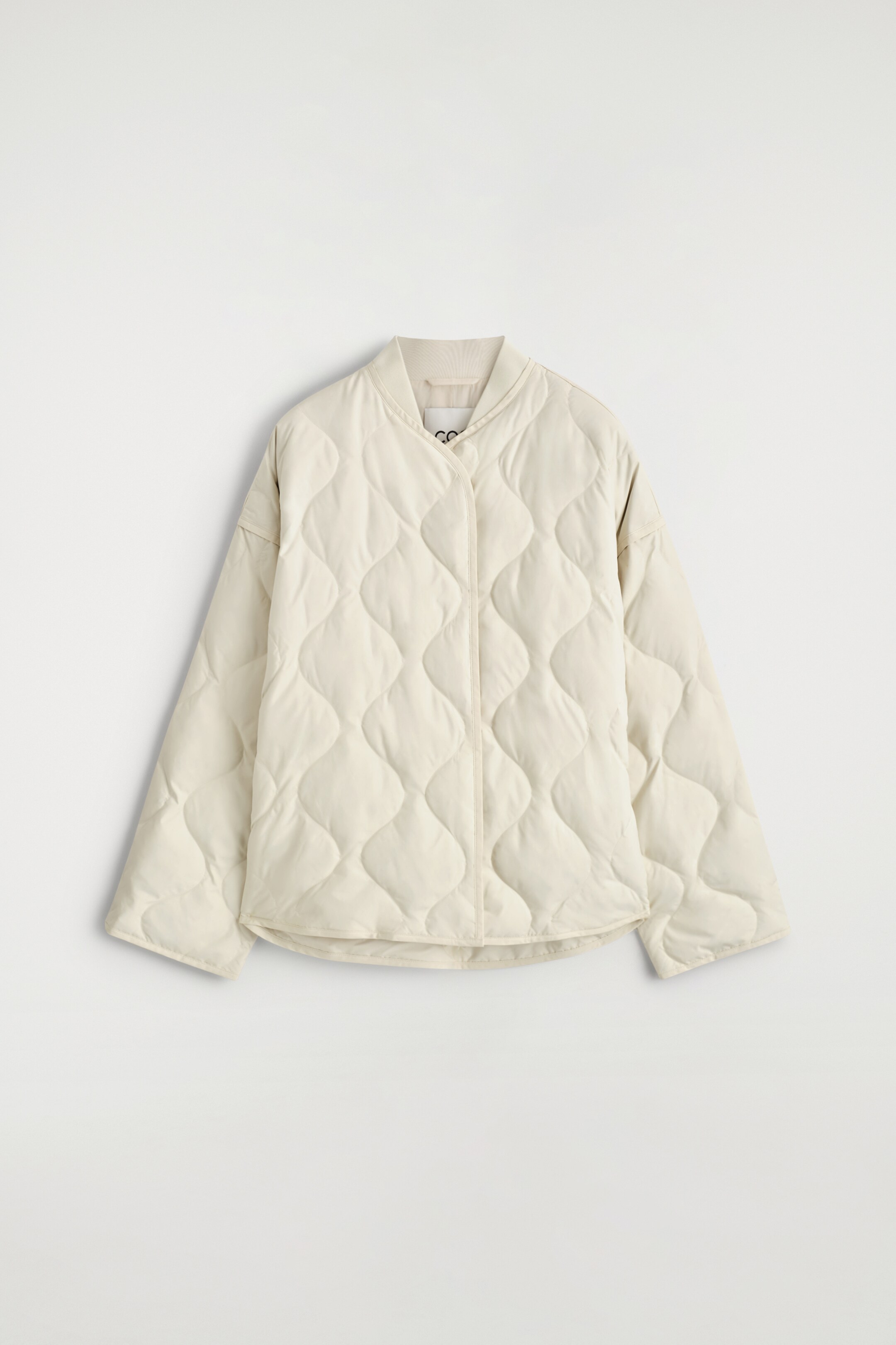 Ver imagem maior: Um casaco acolchoado oversized de cor creme está virado para a frente, apresentando uma gola canelada e um padrão acolchoado em forma de diamante por todo o lado. O seu fecho frontal é uma carcela oculta.