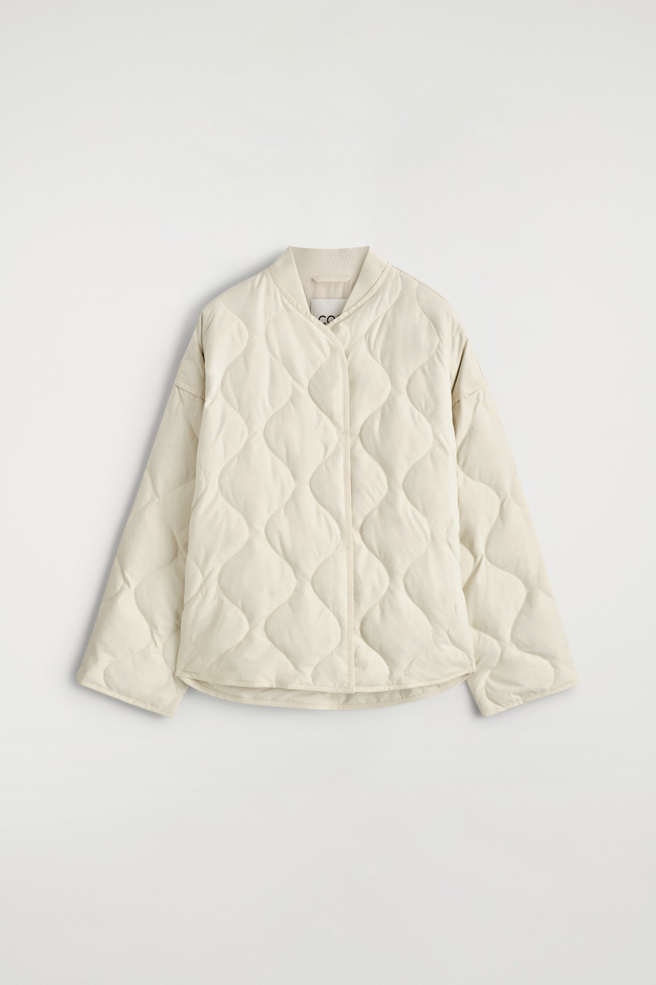 Grösseres Bild ansehen: Eine cremefarbene, übergroße gesteppte Jacke ist nach vorne gerichtet und verfügt über einen gerippten Kragen und ein durchgehendes Rautenmuster. Der Verschluss an der Vorderseite ist eine verdeckte Knopfleiste.