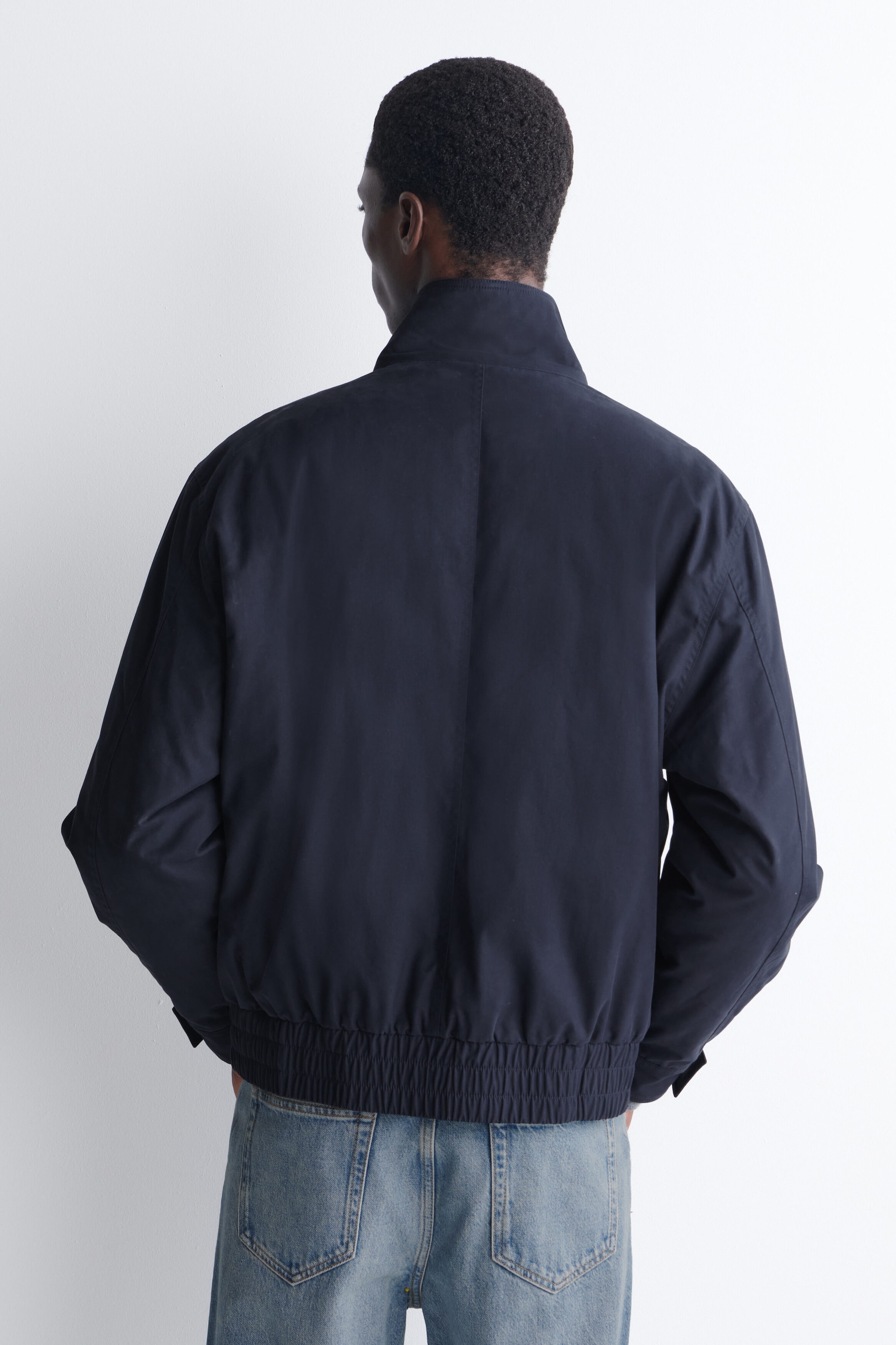 Une veste aviateur rembourrée bleu marine foncé à col cheminée est portée par le mannequin, présenté de dos. La veste présente un col cheminée haut, une couture verticale centrale et un large ourlet élastique froncé, stylisé avec un jean bleu pâle.