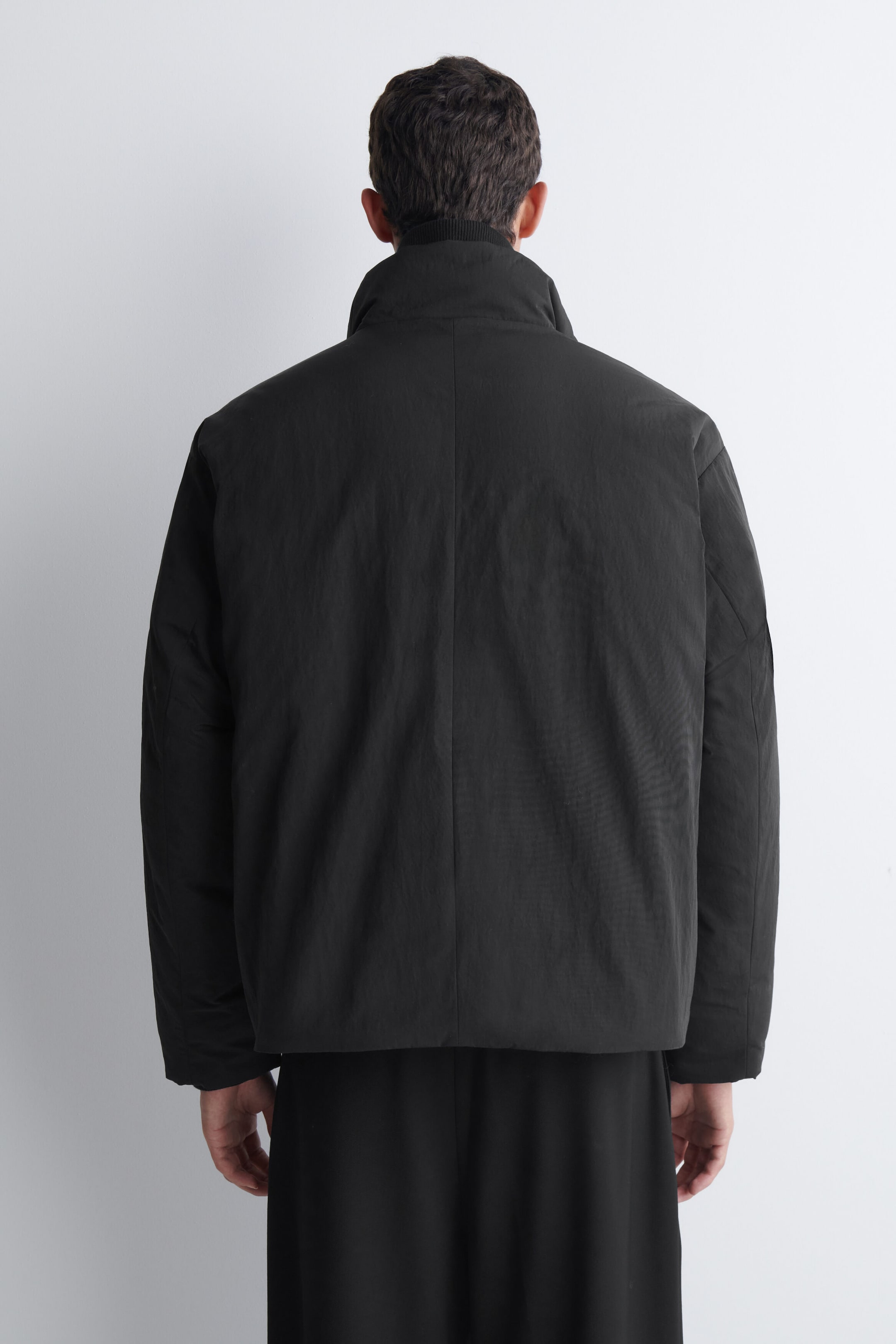 A padded funnel-neck down jacket in matte black is being worn by the model, shown from behind. The jacket features a high, structured collar, a vertical centre-back seam, and a relaxed fit. A padded funnel-neck down jacket in matte black is being worn by the model, shown from behind. The jacket features a high, structured collar, a vertical centre-back seam, and a relaxed fit.